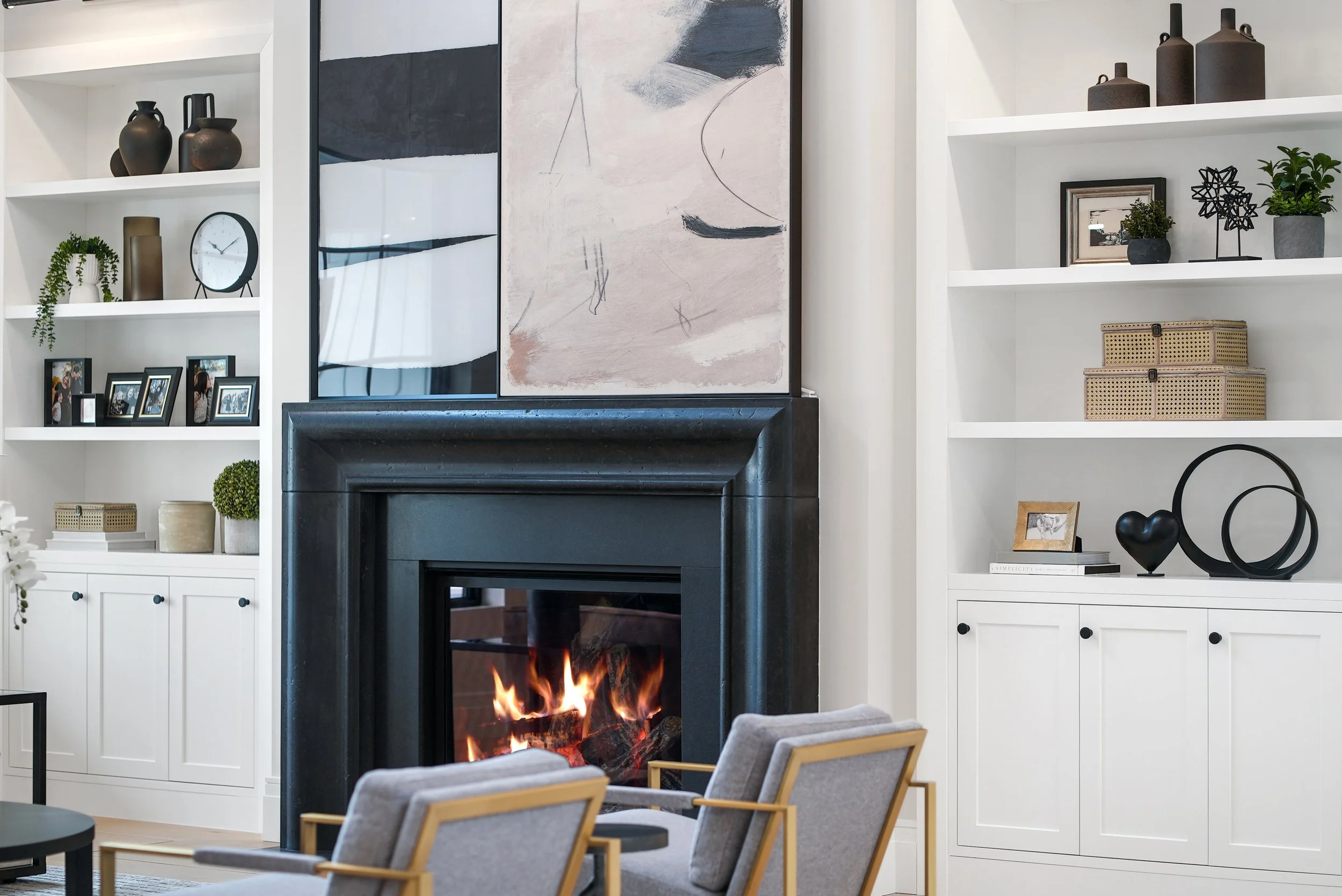 Modern living room with white built-in shelves, black fireplace with a fire, gray armchairs, abstract artwork, and decorative items.