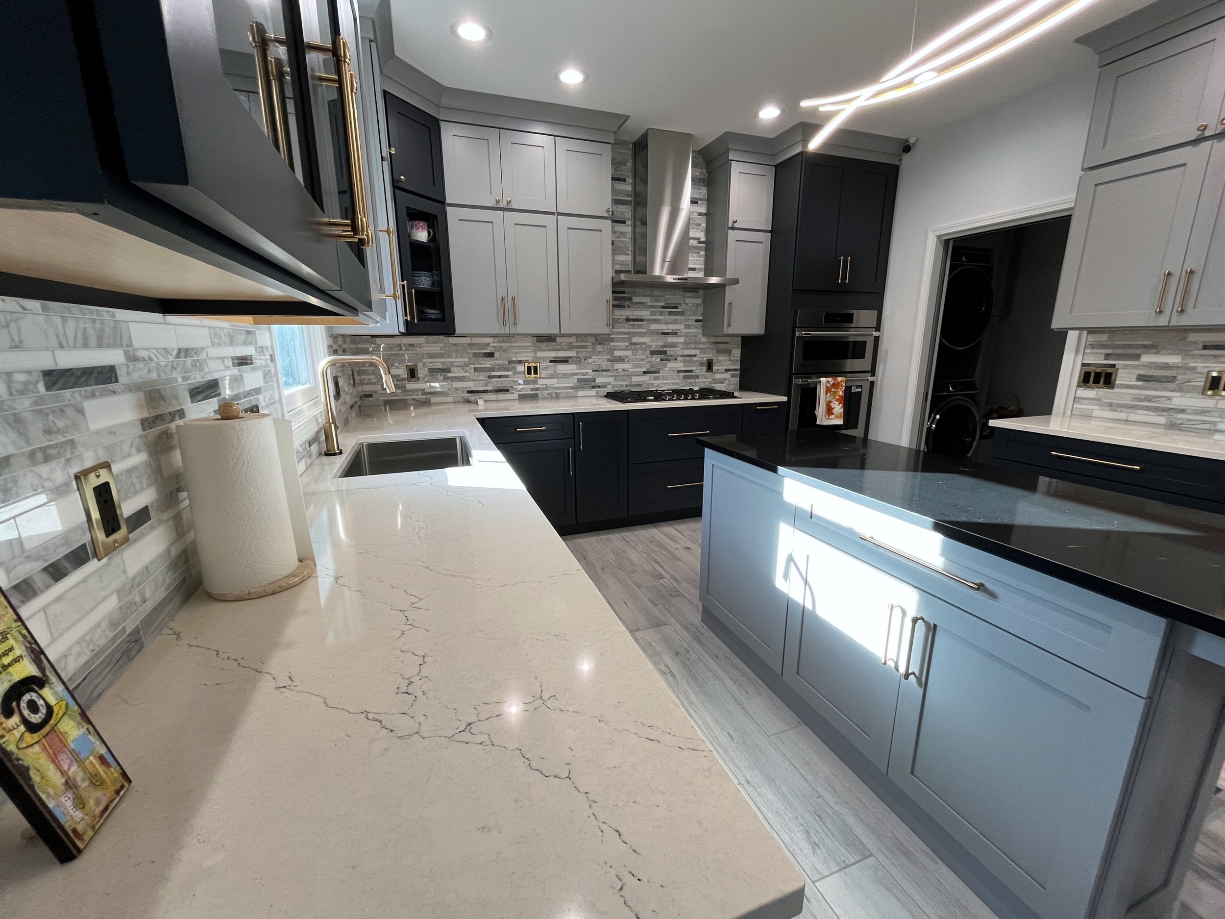 Modern kitchen with gray and black cabinets, marble countertops, stainless steel appliances, tile backsplash, large island, and a washer and dryer in a closet. We specialize in Granite, Quartz, and Marble Countertops. Free Quotes available. 