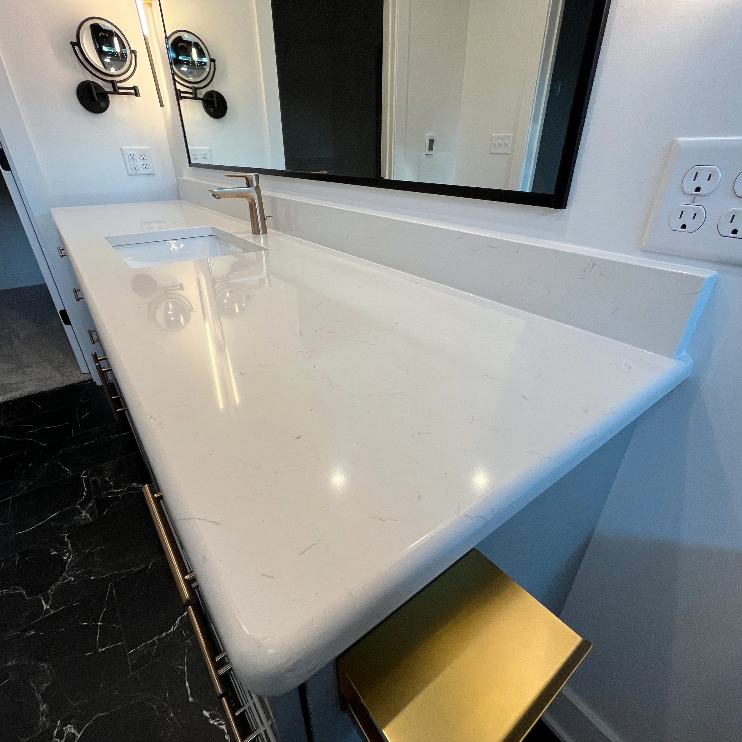 Modern bathroom vanity with a white countertop, a metallic faucet, a large wall mirror, and black marble flooring. We specialize in Granite, Quartz, and Marble Countertops. Free Quotes available. Custom Countertops. Northwest Indiana, Chicagoland