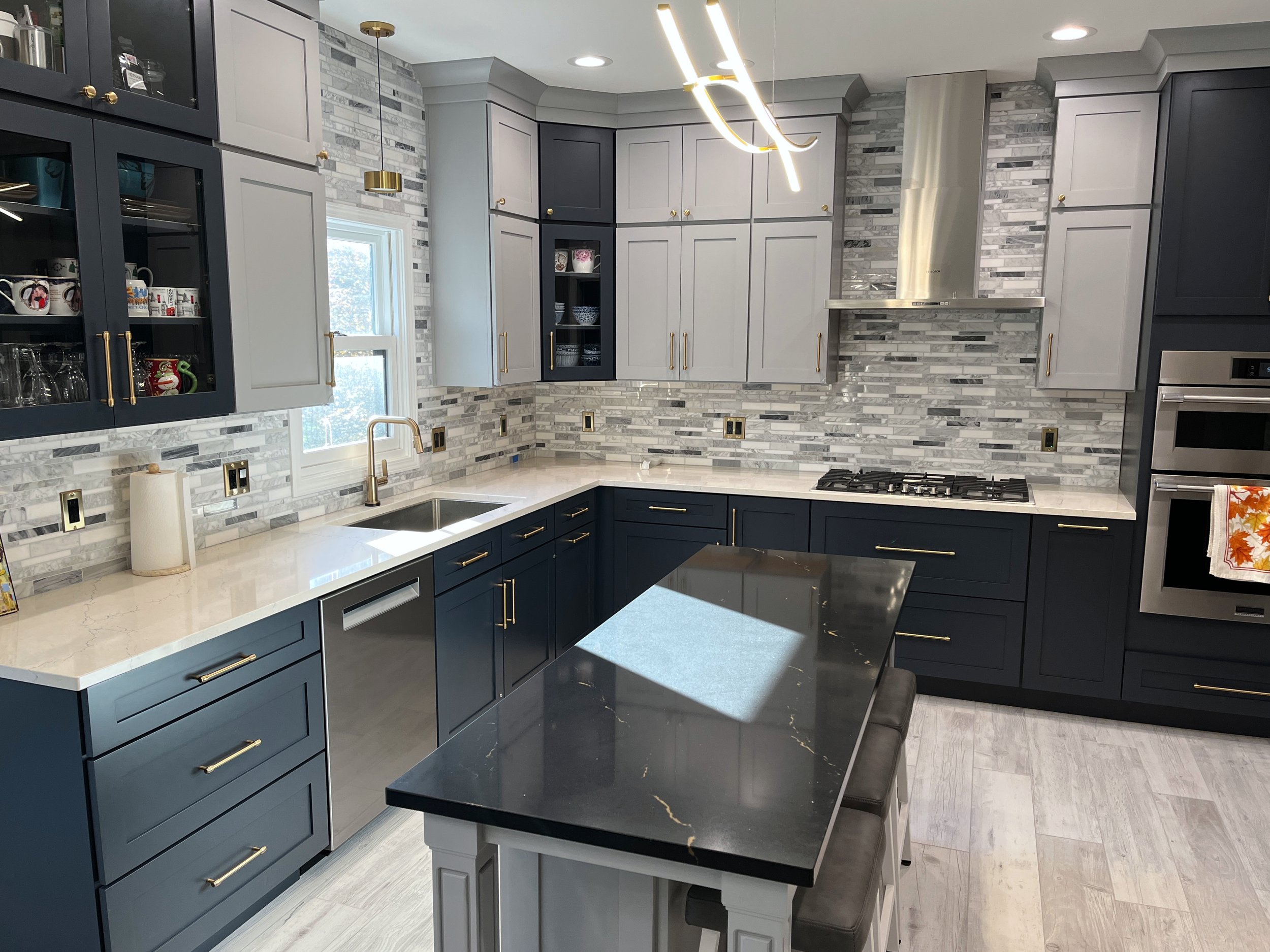 Modern kitchen with gray and navy cabinets, white marble countertops, and a gray and white tile backsplash. Gold handles and fixtures, including a gold pendant light and faucet. The kitchen features a window and stainless steel appliances, with a bla