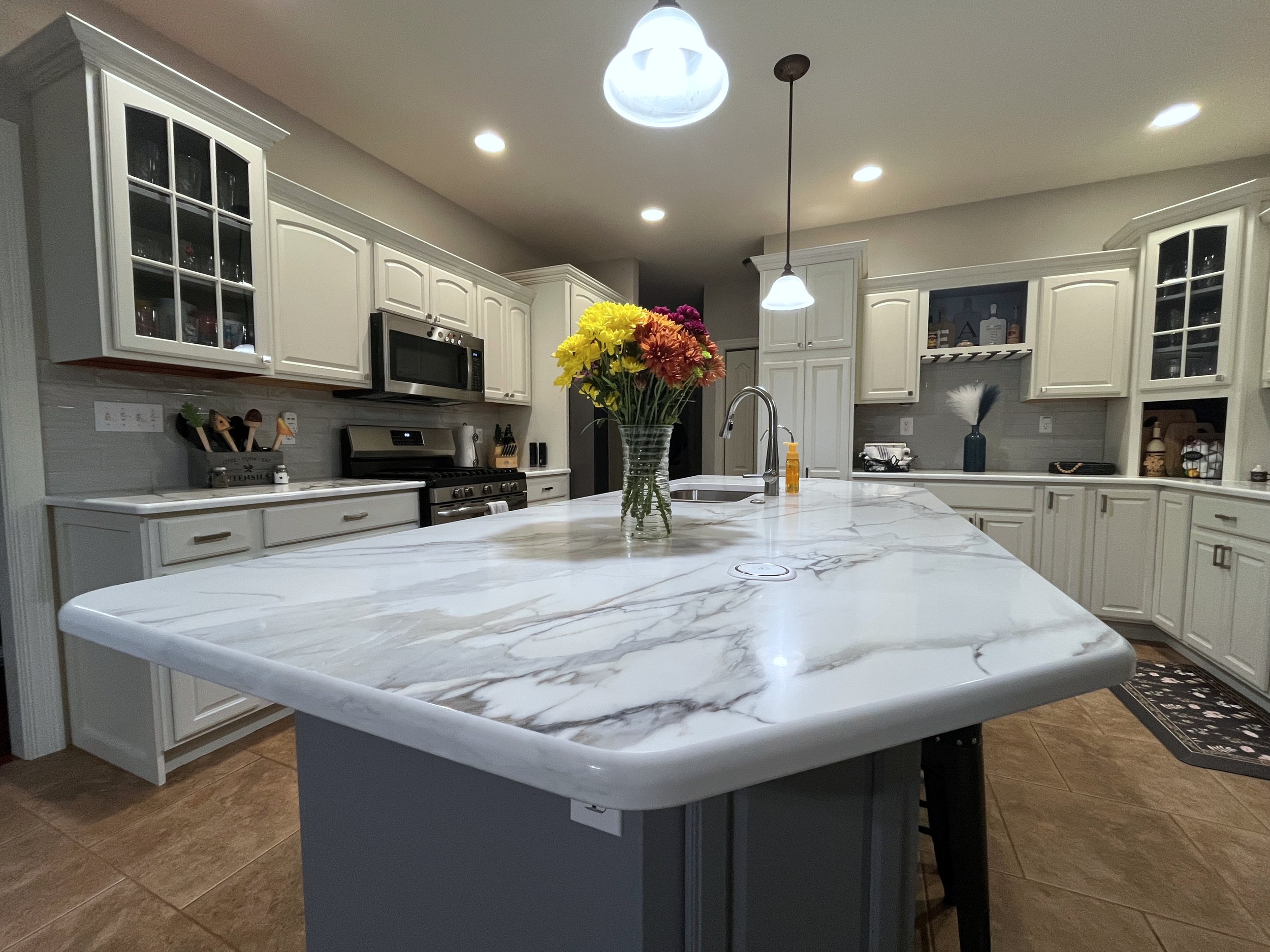 A modern kitchen with white cabinets and granite countertops. A bowl of colorful flowers sits on a large central island. The kitchen has stainless steel appliances, including a microwave and range. There are pendant lights and recessed lighting in the ceiling, and decorative items on the shelves and counters.