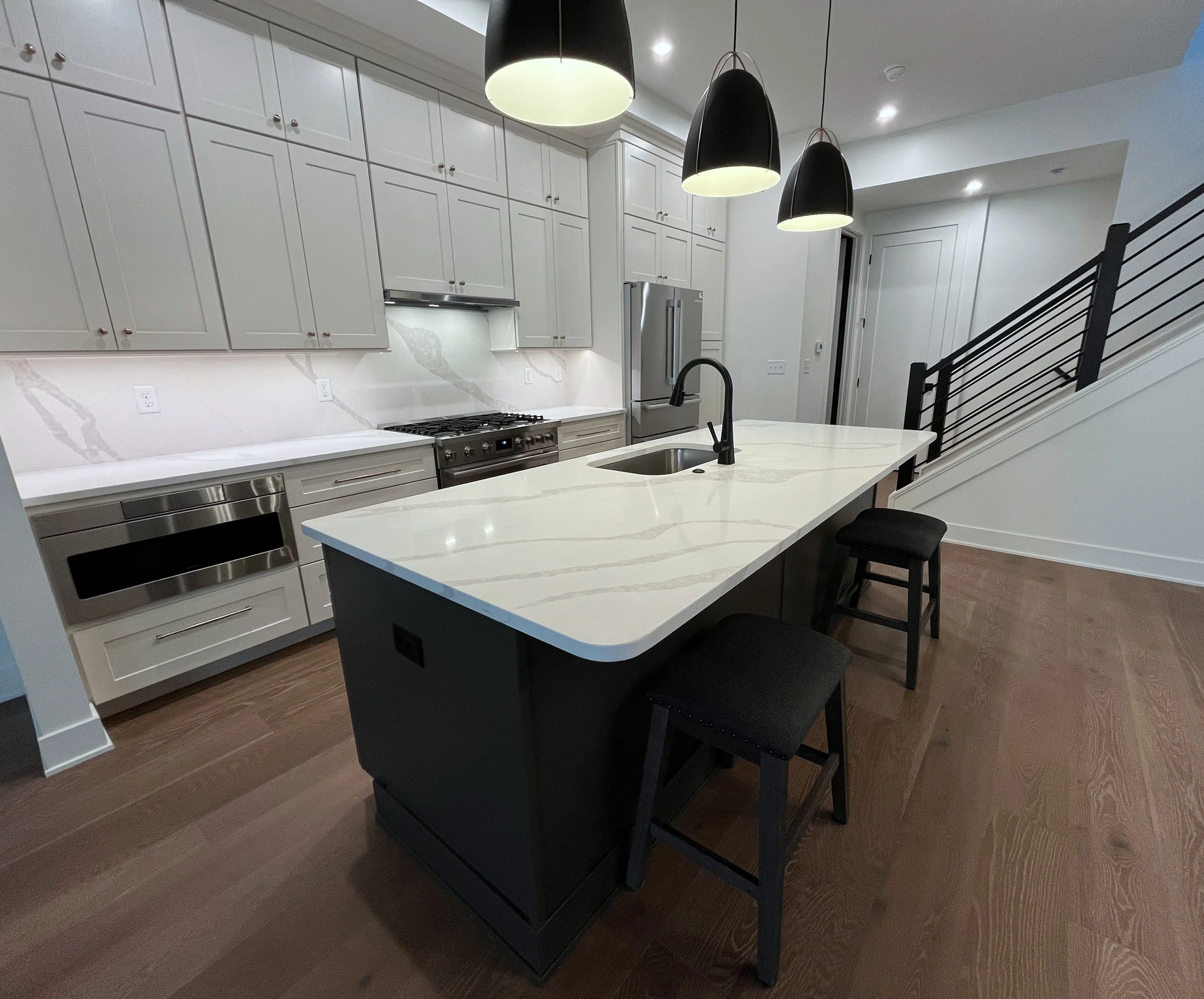 Modern kitchen with white cabinets, stainless steel appliances, black pendant lights, and a white marble island with a black faucet and two black chairs. We specialize in Granite, Quartz, and Marble Countertops. Free Quotes available. Custom Counters