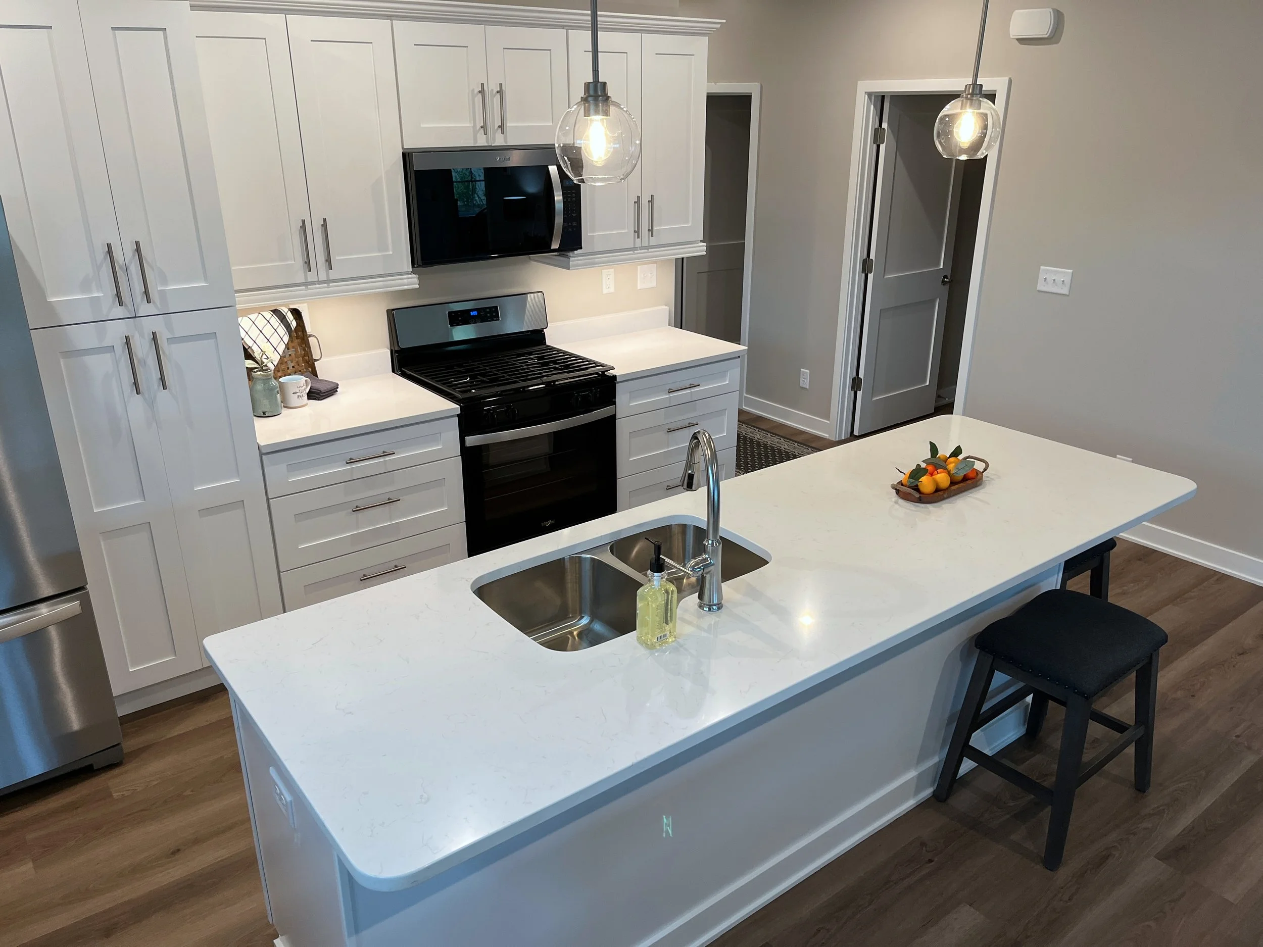 Modern kitchen with white cabinets, a black microwave, oven, and a white countertop island with a bowl of fruit, pendant lights, and hardwood floors. We specialize in Granite, Quartz, and Marble Countertops. Free Quotes available. Custom Countertops.