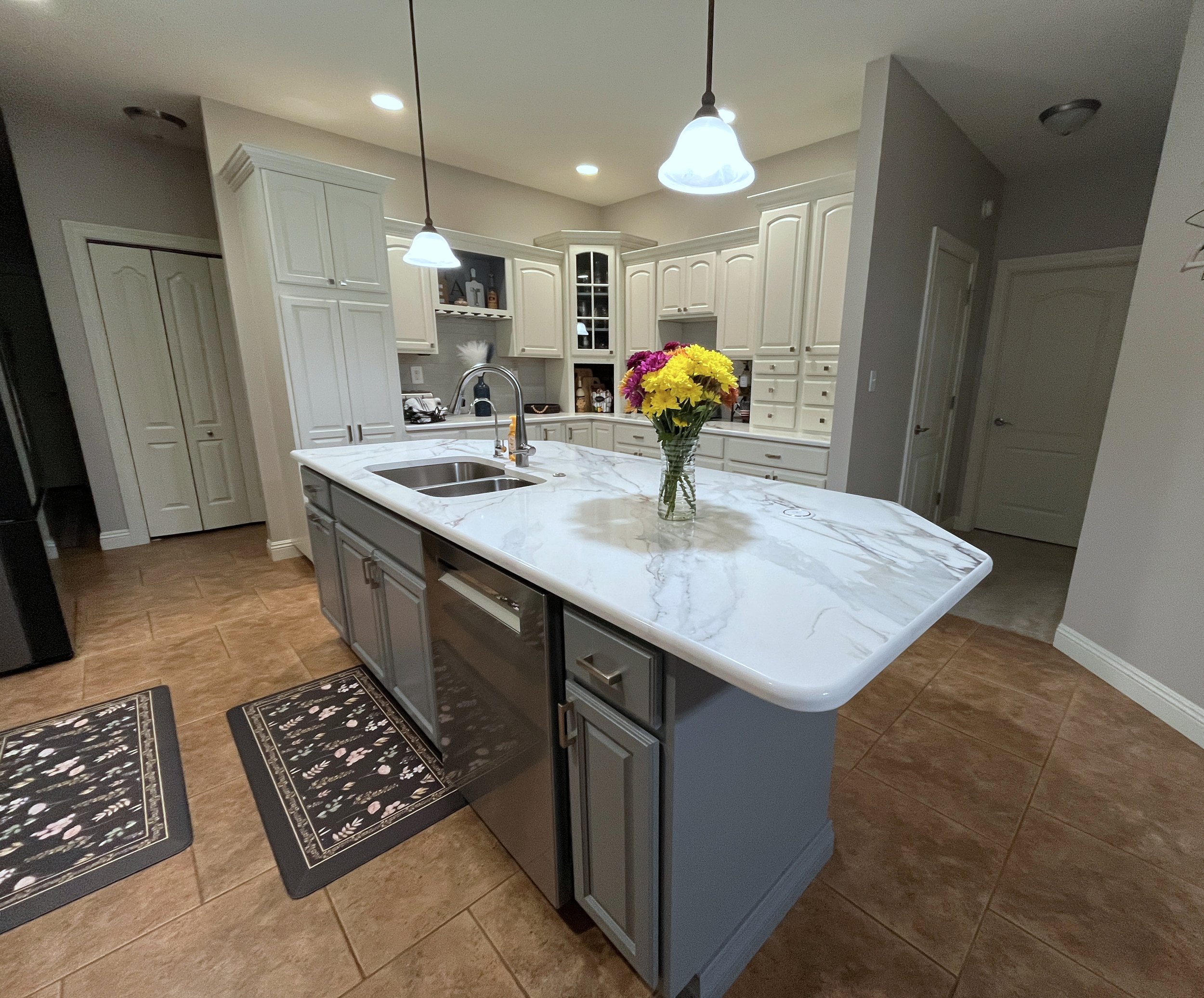 A kitchen with white cabinets, a marble-topped island, and a vase of colorful flowers. There are two black and floral rugs on the tiled floor, and pendant lights hanging from the ceiling. We specialize in Granite, Quartz, and Marble Countertops.