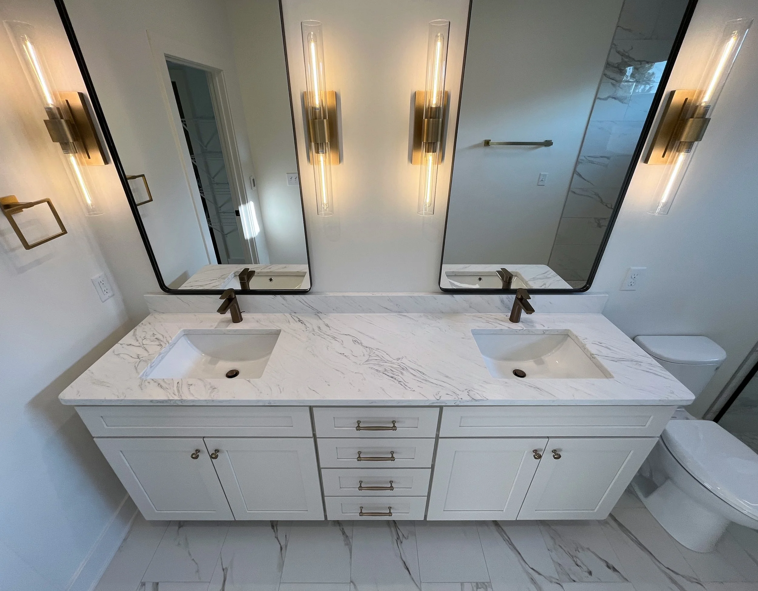 Bathroom vanity with double sinks, white marble countertop, gold hardware, large mirror, modern light fixtures, and neutral wall colors. We specialize in Granite, Quartz, and Marble Countertops. Free Quotes available. Custom Countertops. Northwest.