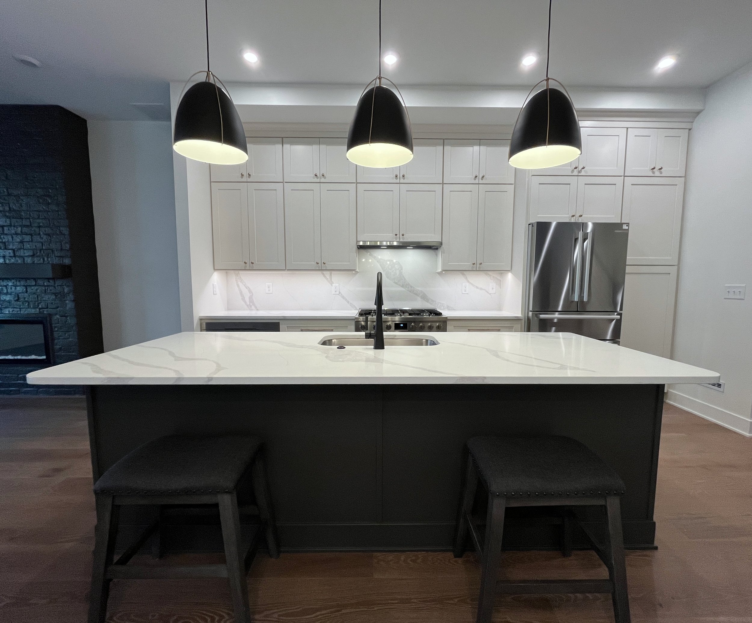 Modern kitchen with white cabinets, a marble countertop, a stainless steel refrigerator, and three black pendant lights hanging above the island. We specialize in Granite, Quartz, and Marble Countertops. Free Quotes available. Custom Countertops.