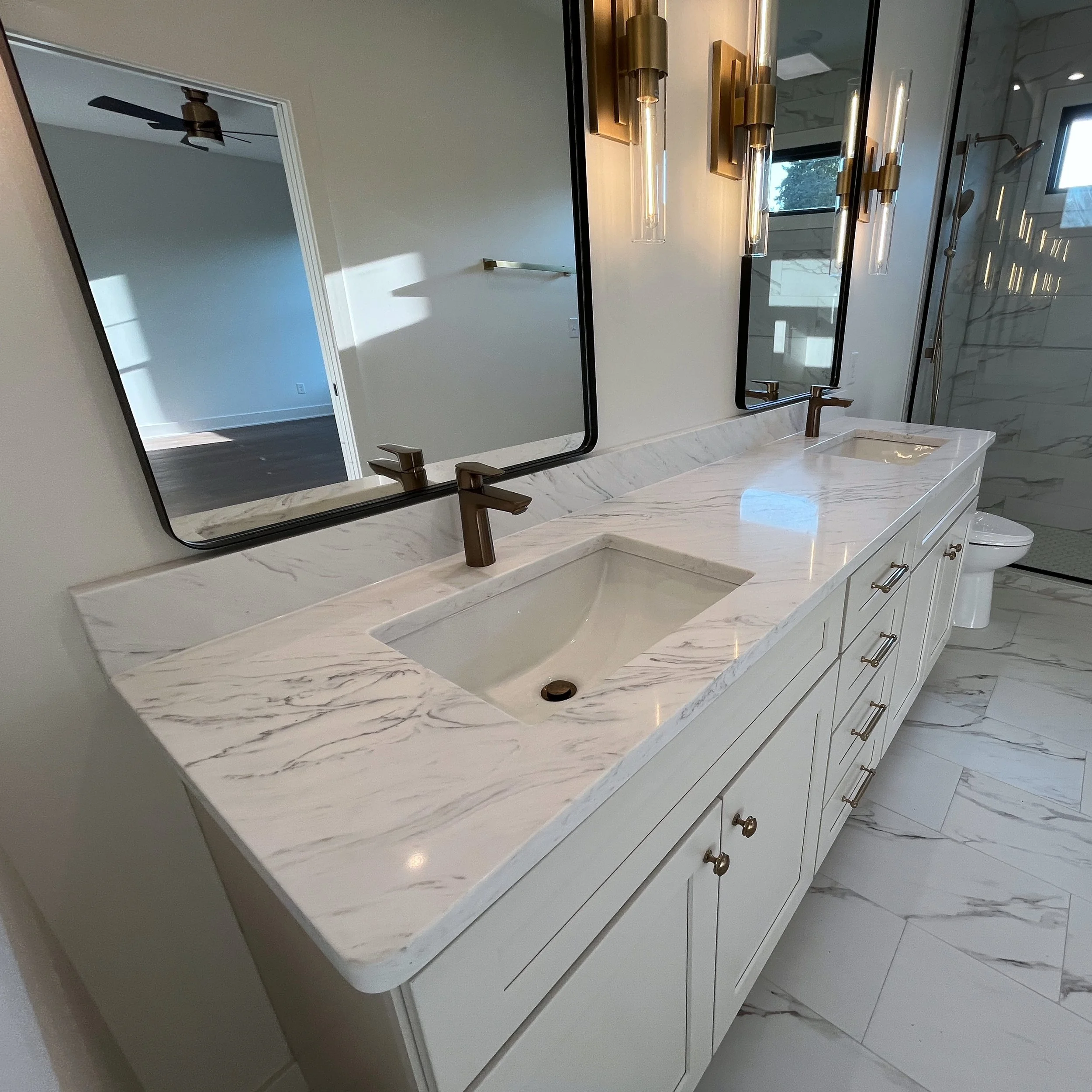 Modern bathroom with a marble countertop, two sinks, large mirror, gold fixtures, and a walk-in shower with glass doors. We specialize in Granite, Quartz, and Marble Countertops. Free Quotes available. Custom Countertops. Northwest Indiana, Chicago.
