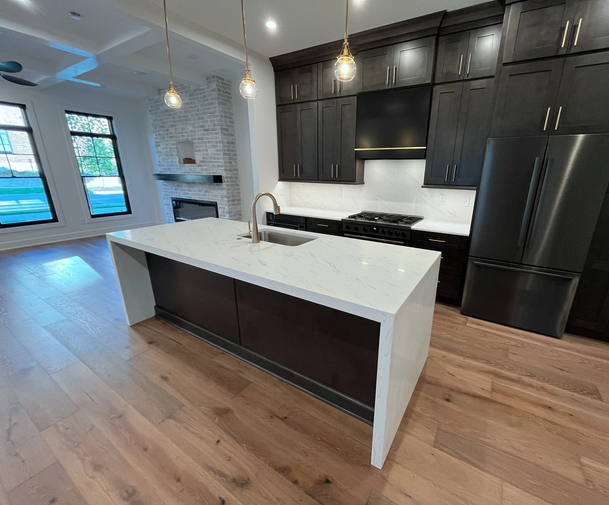 Modern kitchen with dark cabinets, white marble island, stainless steel appliances, and hardwood floors, with large windows overlooking outdoors. We specialize in Granite, Quartz, and Marble Countertops. Free Quotes available. Custom Countertops. 
