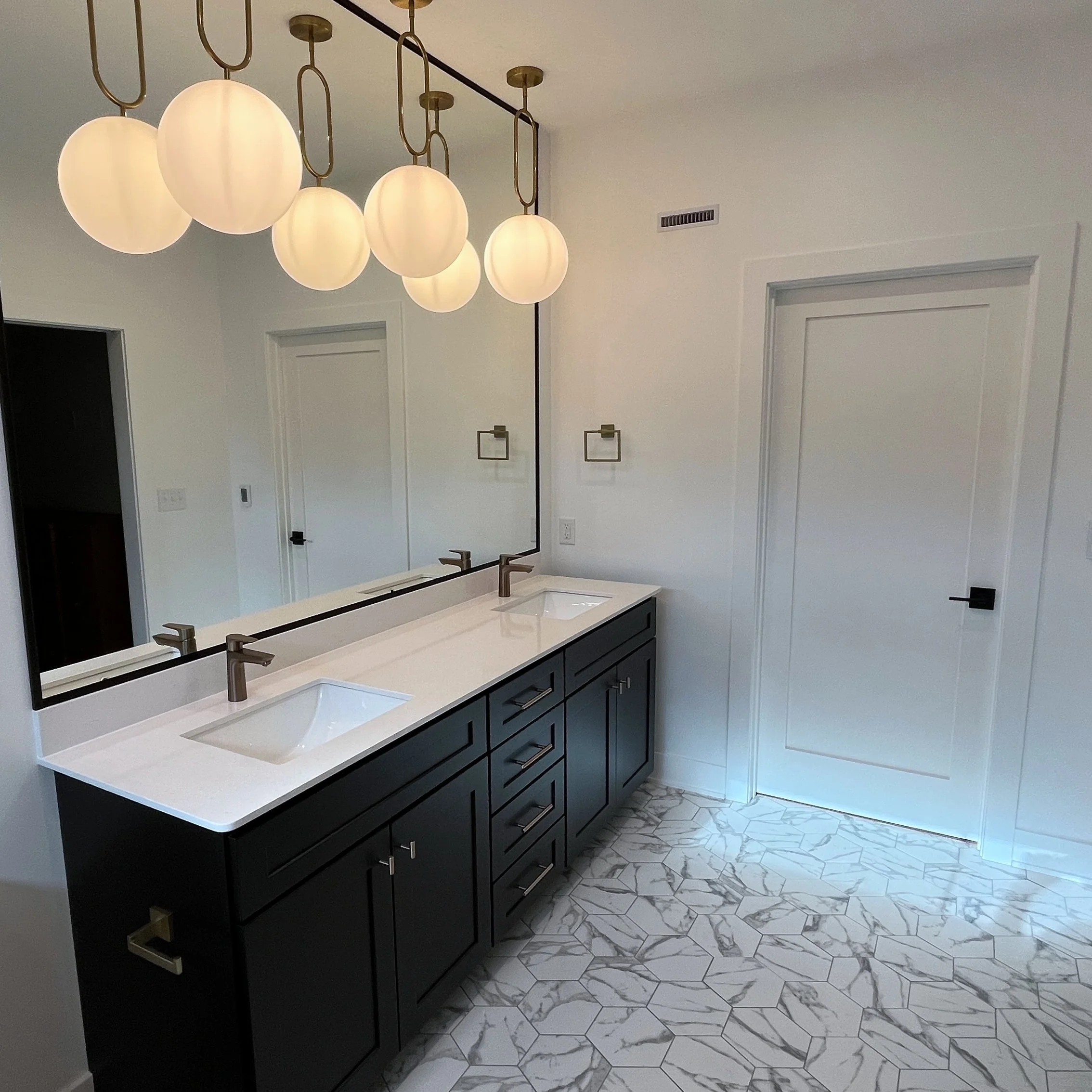 Modern bathroom with white marble hexagon tile flooring, black vanity with gold handles, large mirror, and a white countertop with dual sinks. Overhead, a chandelier with multiple round, frosted glass globes hangs from the ceiling. 