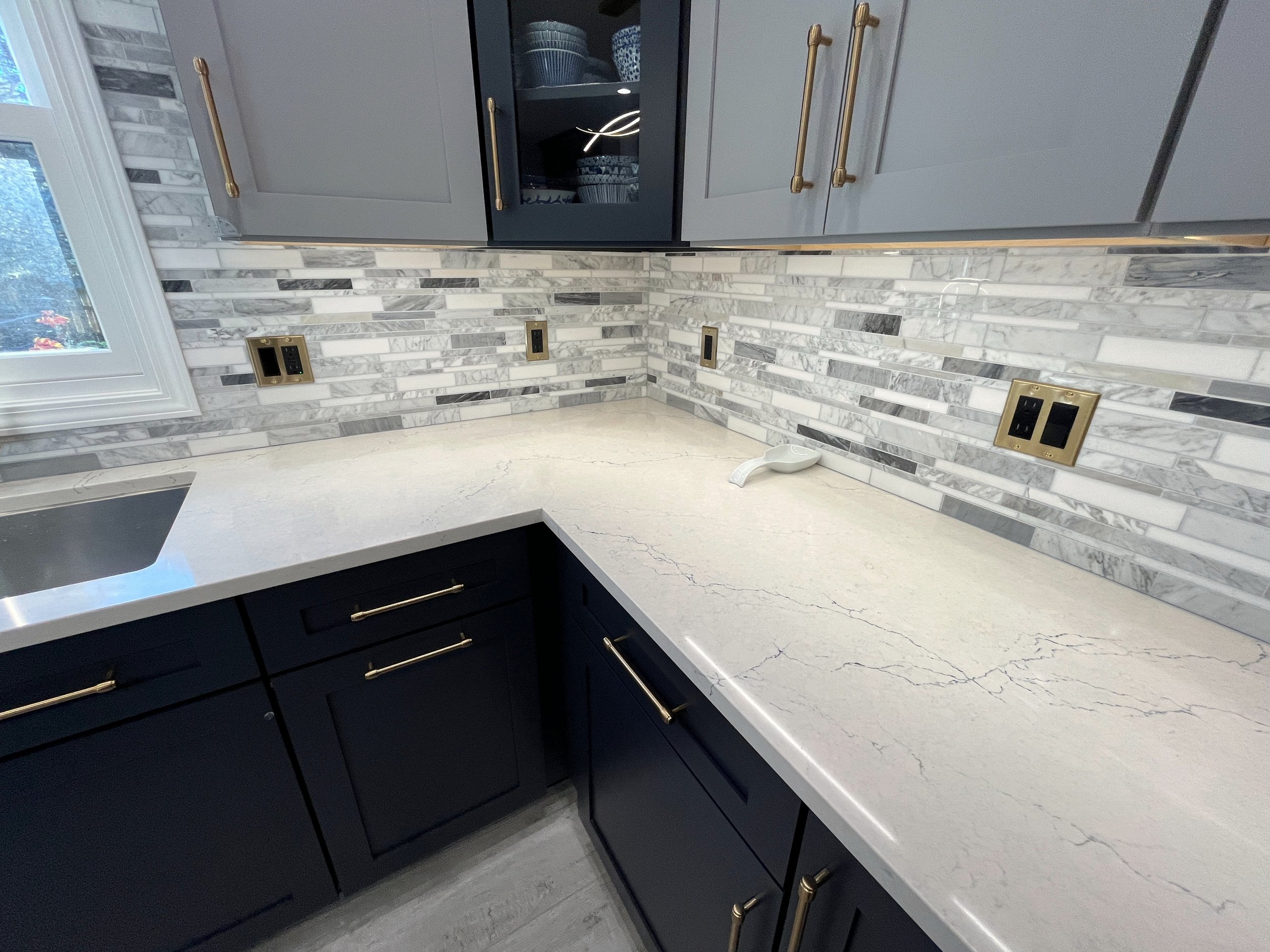 Kitchen with white marble countertops, dark navy cabinets with gold handles, a window on the left, and a backsplash made of white and gray rectangular tiles. There are electrical outlets with gold covers on the backsplash, and a white bowl with a han