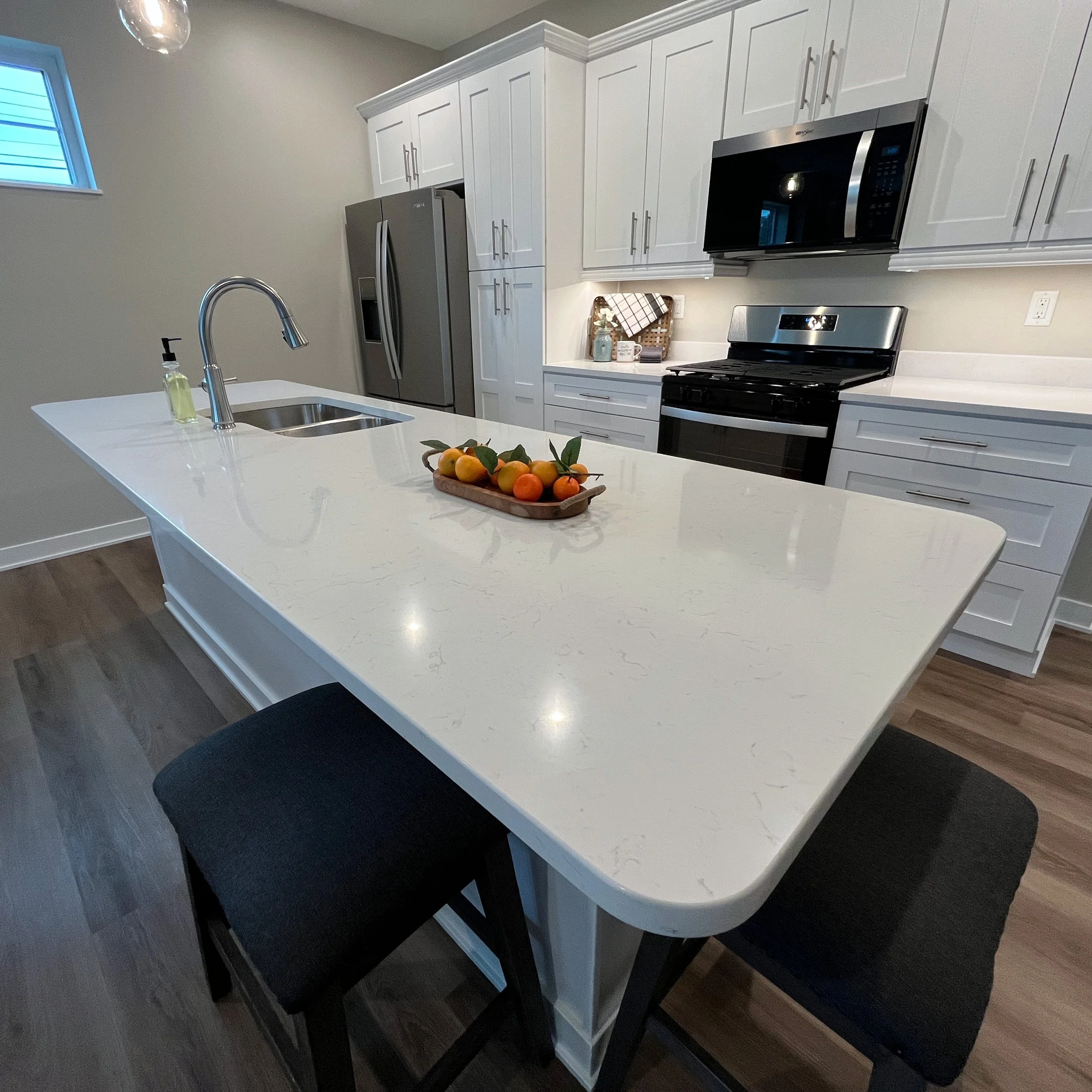 A modern kitchen with white cabinets, stainless steel appliances, a kitchen island with a white countertop, a fruit basket, and a window with blue blinds. We specialize in Granite, Quartz, and Marble Countertops. Free Quotes available. Custom Counter