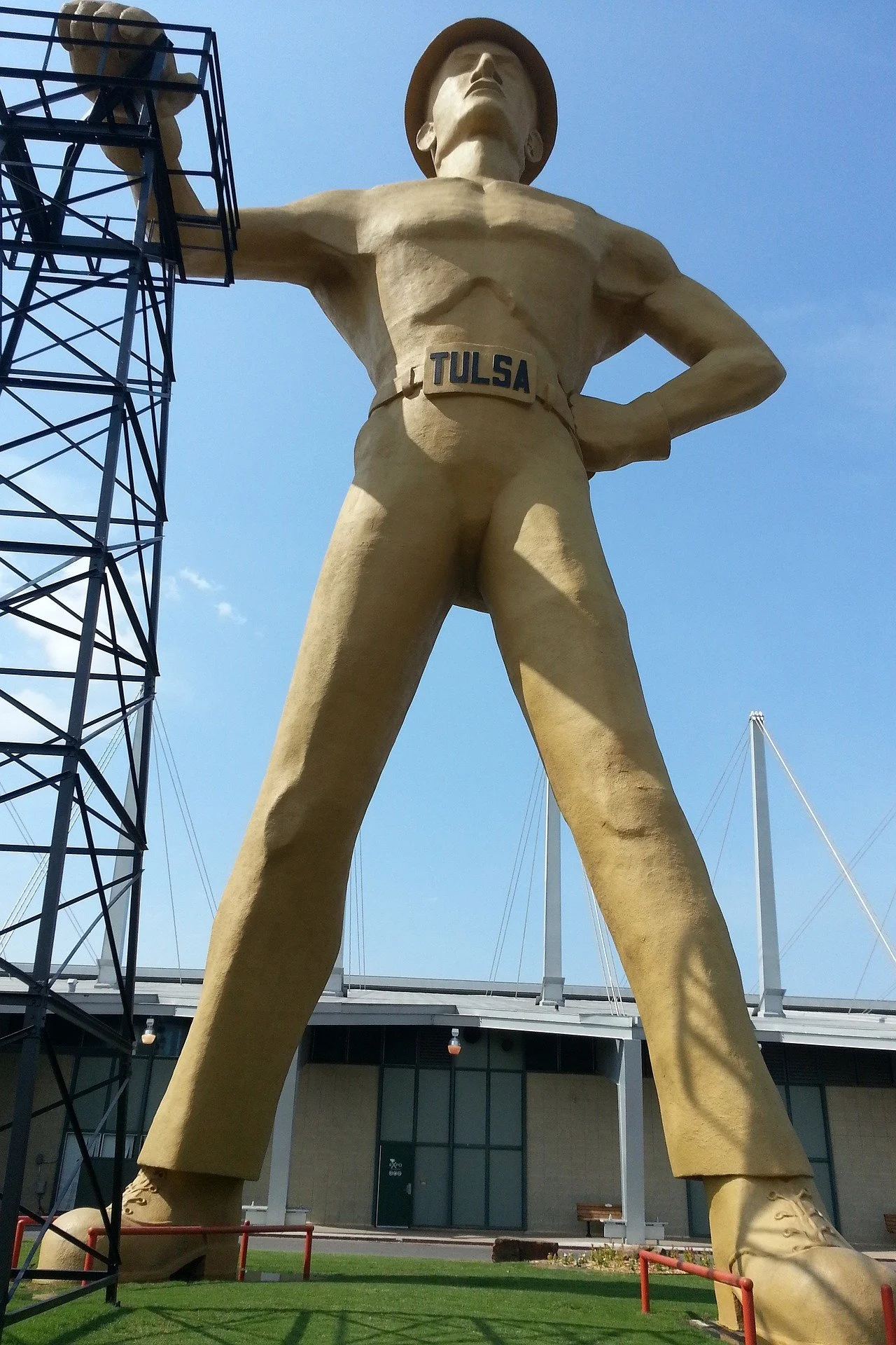 A giant statue of a man in a cowboy outfit with a hat, with the word 'Tulsa' on his belt, standing with one arm raised holding a solar panel, and the other hand on his hip. The statue is outside near a building and a marina, with a blue sky overhead.