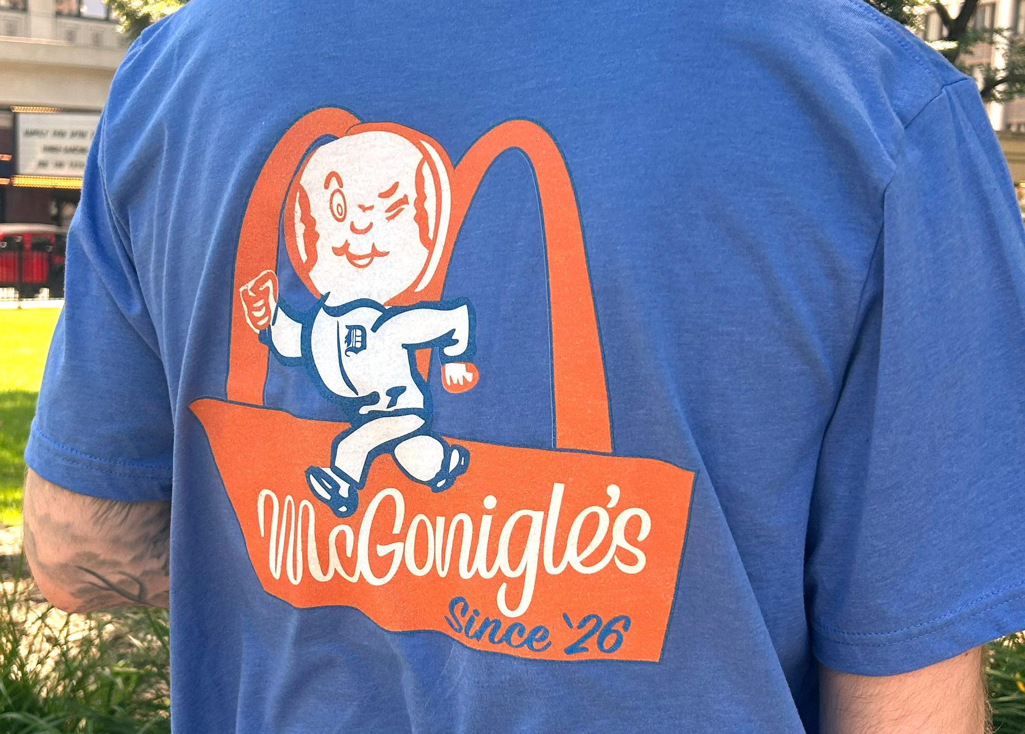 Close-up of a person wearing a blue t-shirt with a cartoon graphic of a smiling baseball, featuring the text 'McGonigle's since '26' in orange and white colors.