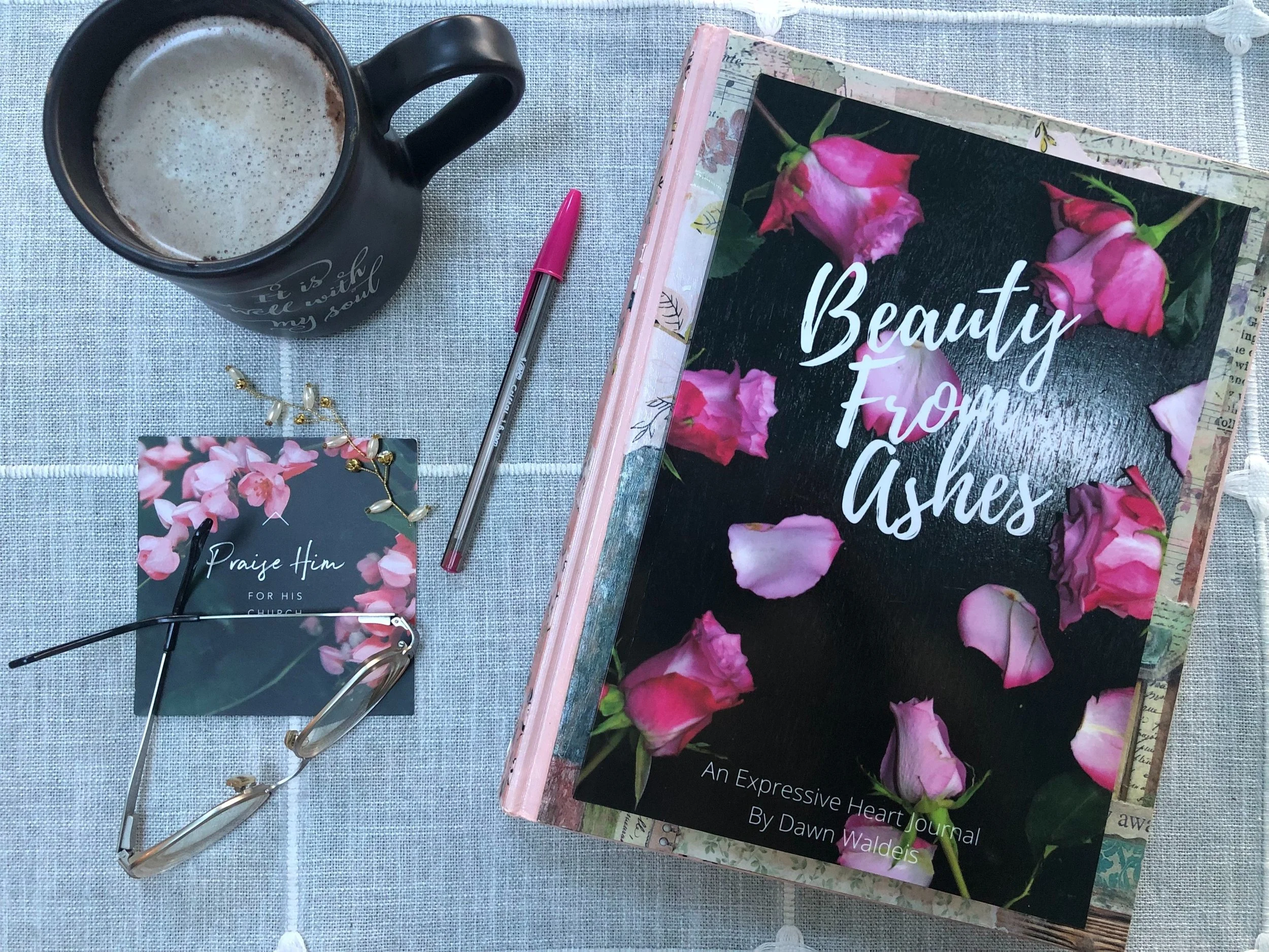A table with a black ceramic mug filled with a frothy beverage, a pink pen, reading glasses, a small card with pink flowers and the words 'Praise Him for His Church,' and a scrapbook titled 'Beauty from Ashes' containing pink roses on the cover.
