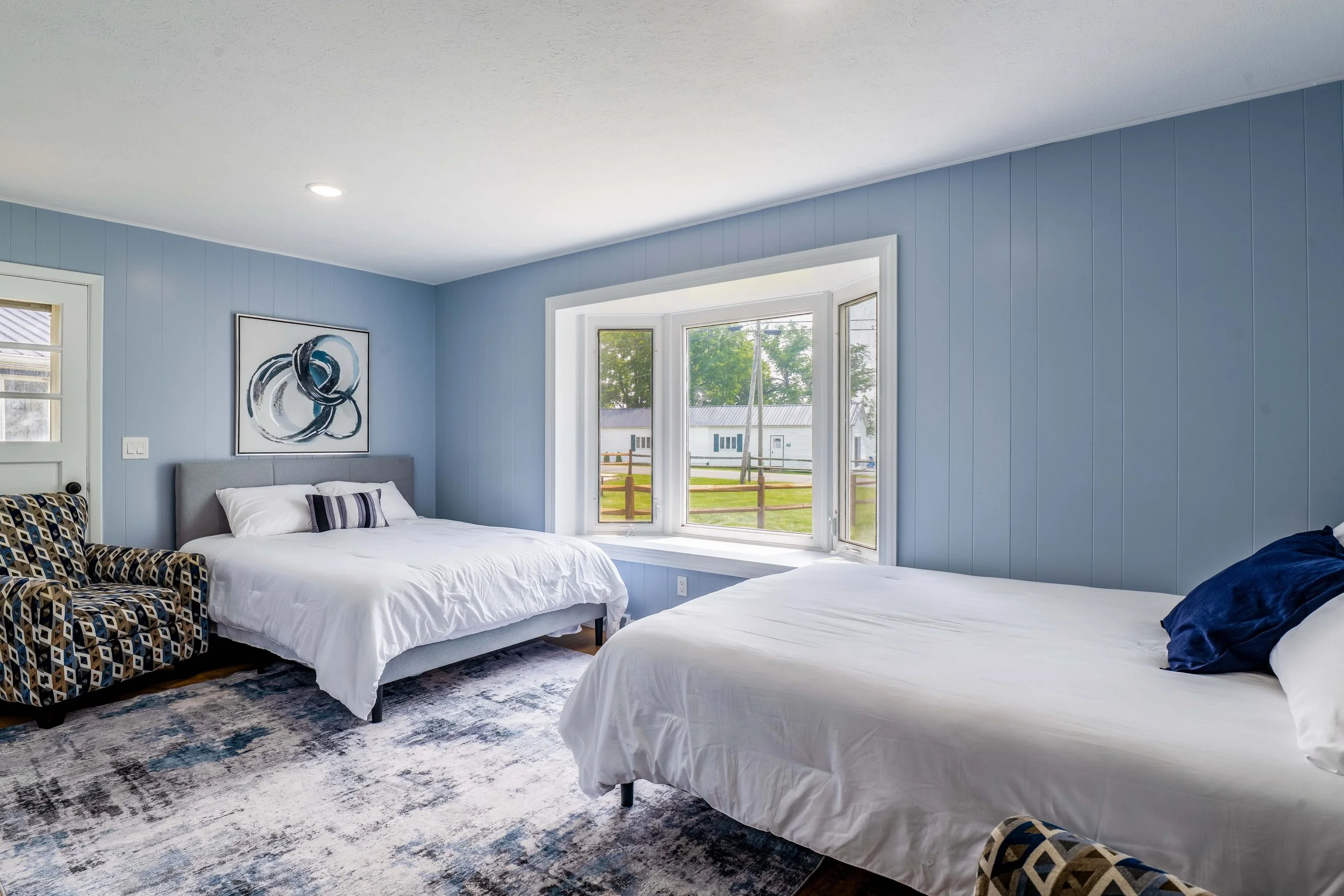 A bedroom with light blue painted walls, two white beds, a patterned armchair, a large window with a view of trees and a building outside, a modern abstract wall art, and a patterned area rug.