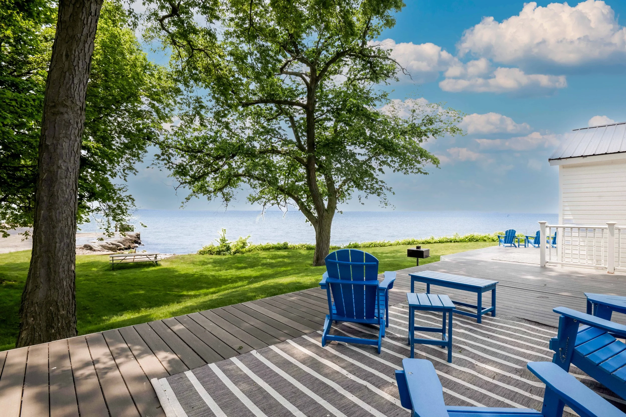 A backyard deck with blue Adirondack chairs, facing a grassy area with trees, a rocky shoreline, and a body of water in the distance under a partly cloudy sky.