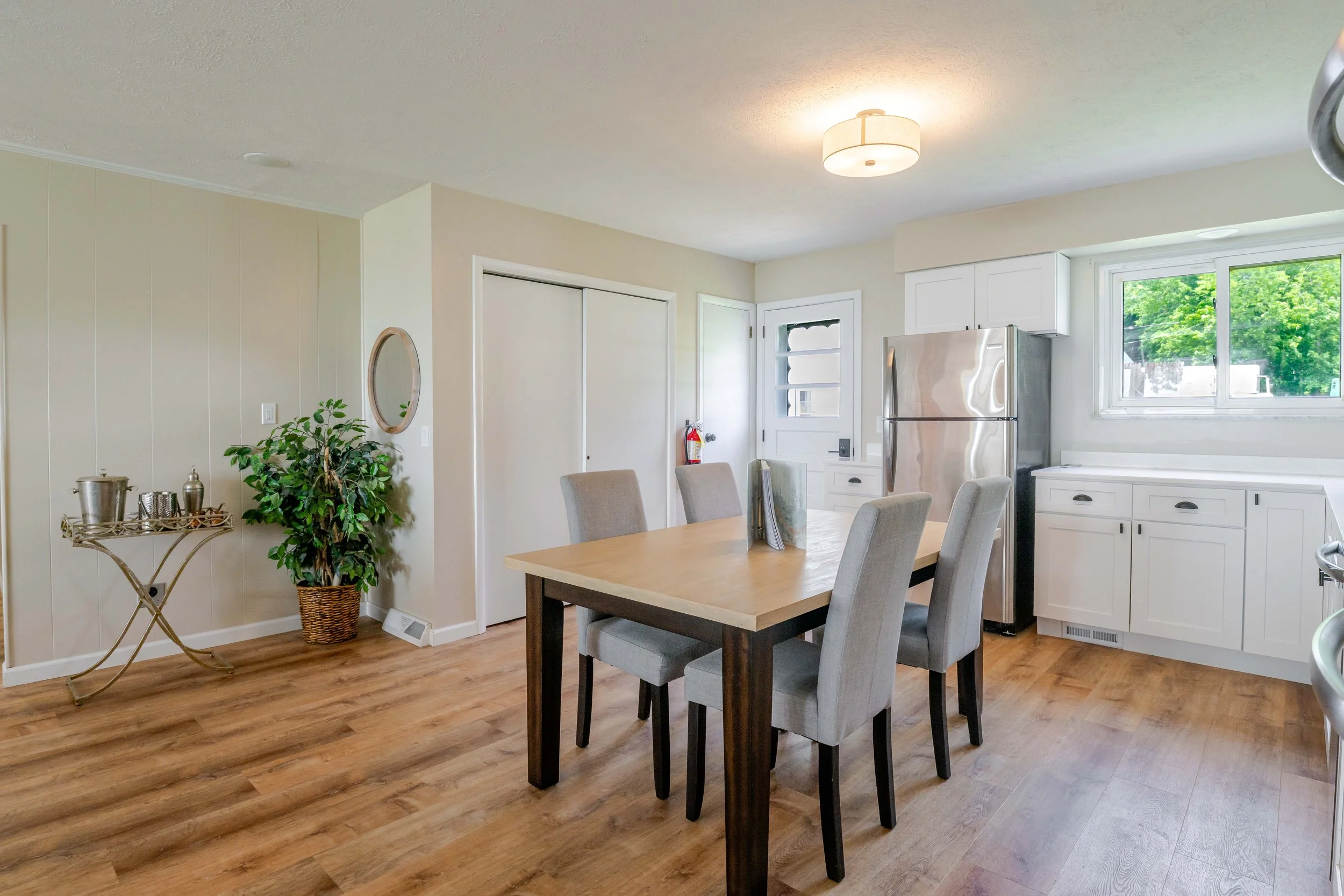A cozy kitchen and dining area with a wooden table and gray upholstered chairs, a stainless steel refrigerator, white cabinets, a window with green trees outside, a potted plant, a decorative mirror, and wood flooring.
