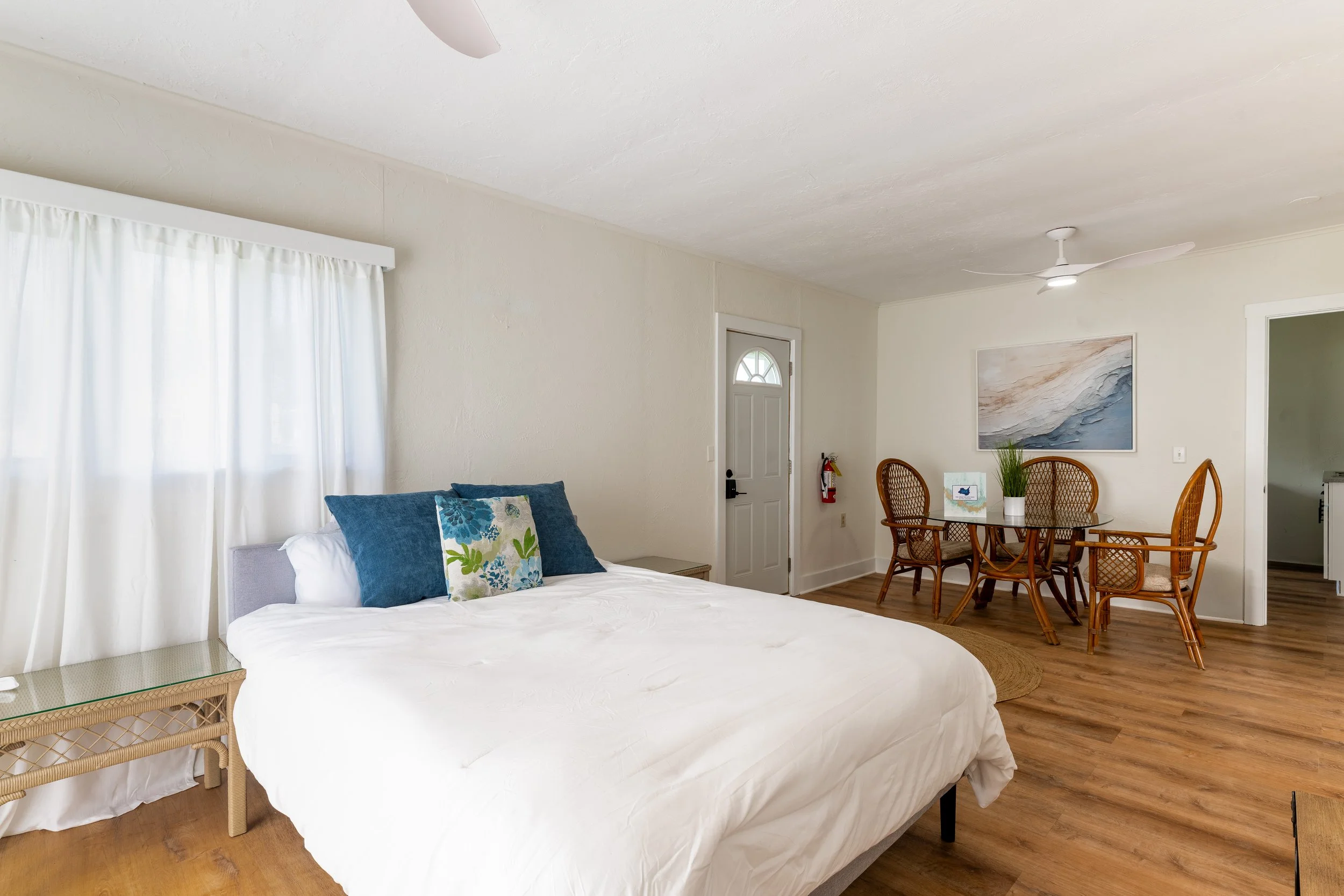 A bedroom with a bed that has a white bedspread and decorative pillows, beside a wicker nightstand, with a window with white curtains, and a wooden dining table with wicker chairs, wall art, a ceiling fan, and a door in the background.
