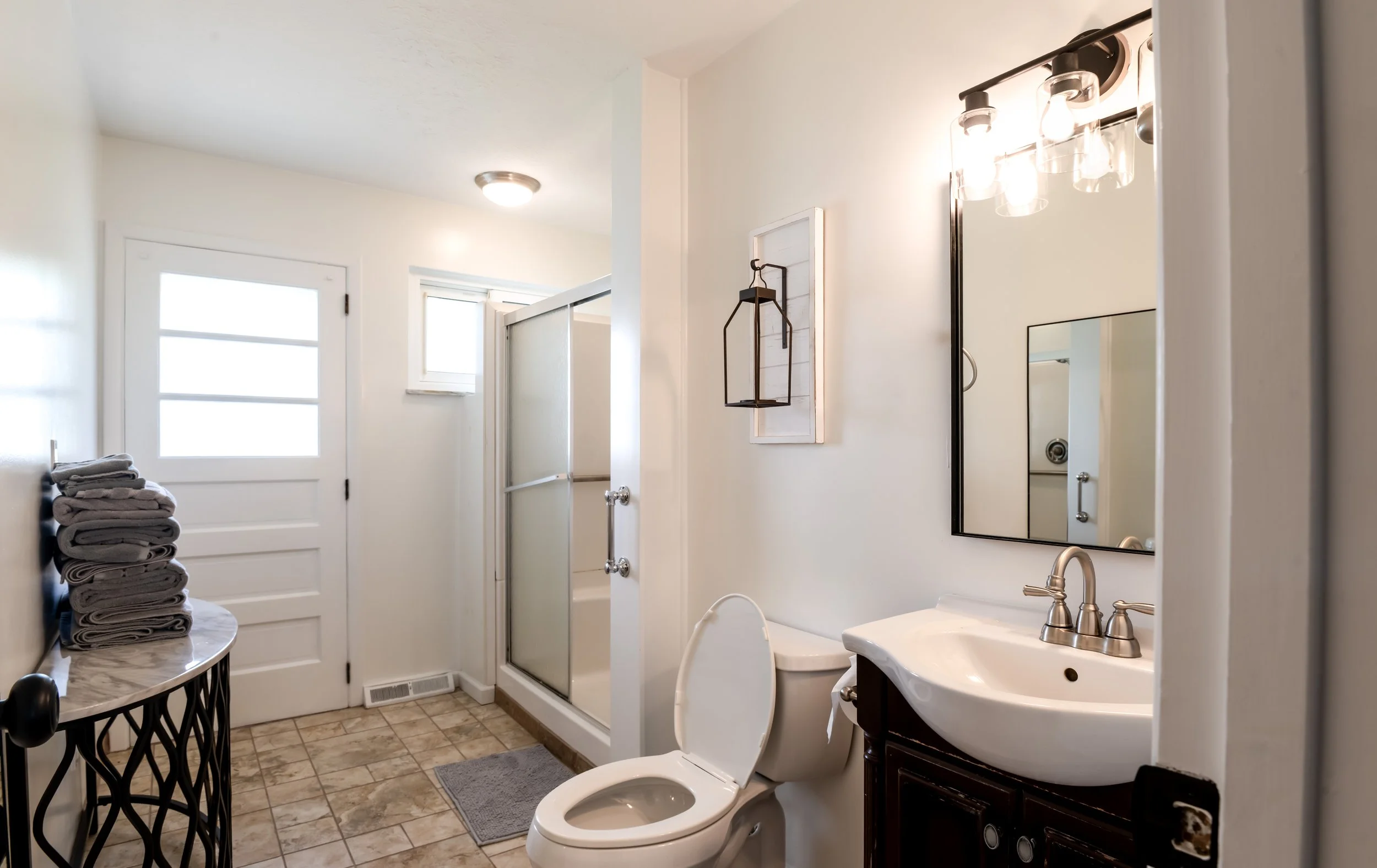 A bathroom with a white door and windows, a stack of folded towels on a side table, a mirror above a beige vanity with a dark wood cabinet, a toilet, a small gray rug, and a shower with a sliding door. The walls are painted white and the floor is til