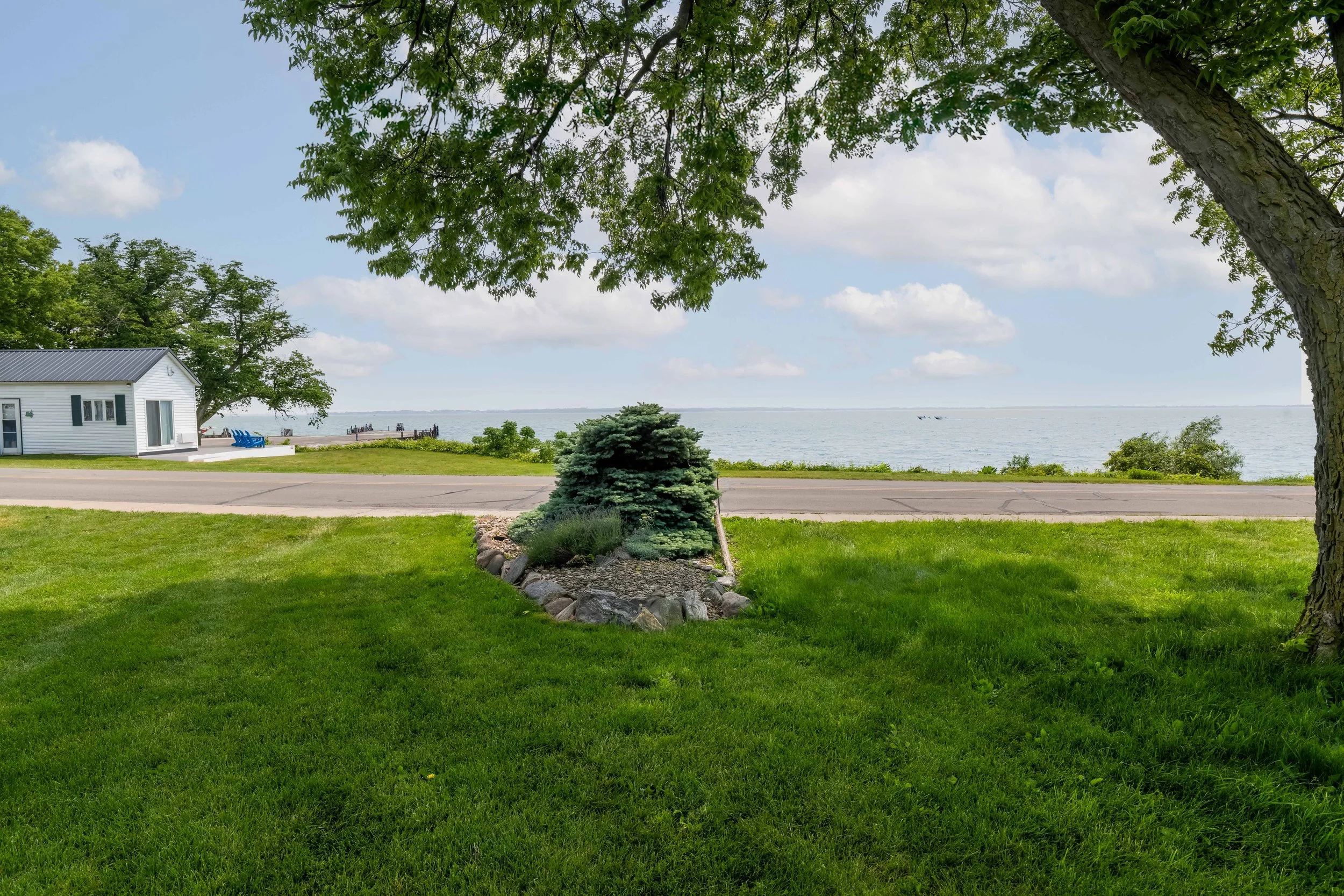 View of a lawn and garden with a white house, a water view in the background, and green trees providing shade.