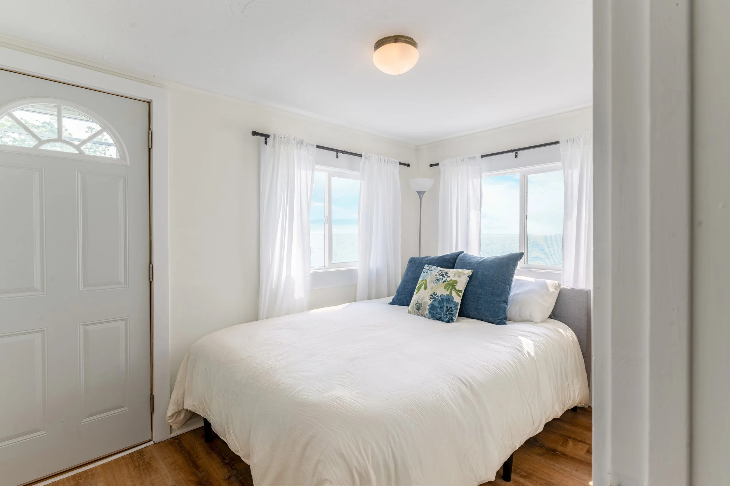 A cozy bedroom with white walls, a white bed with floral and navy pillows, large windows with white curtains, a floor lamp, and a ceiling light.