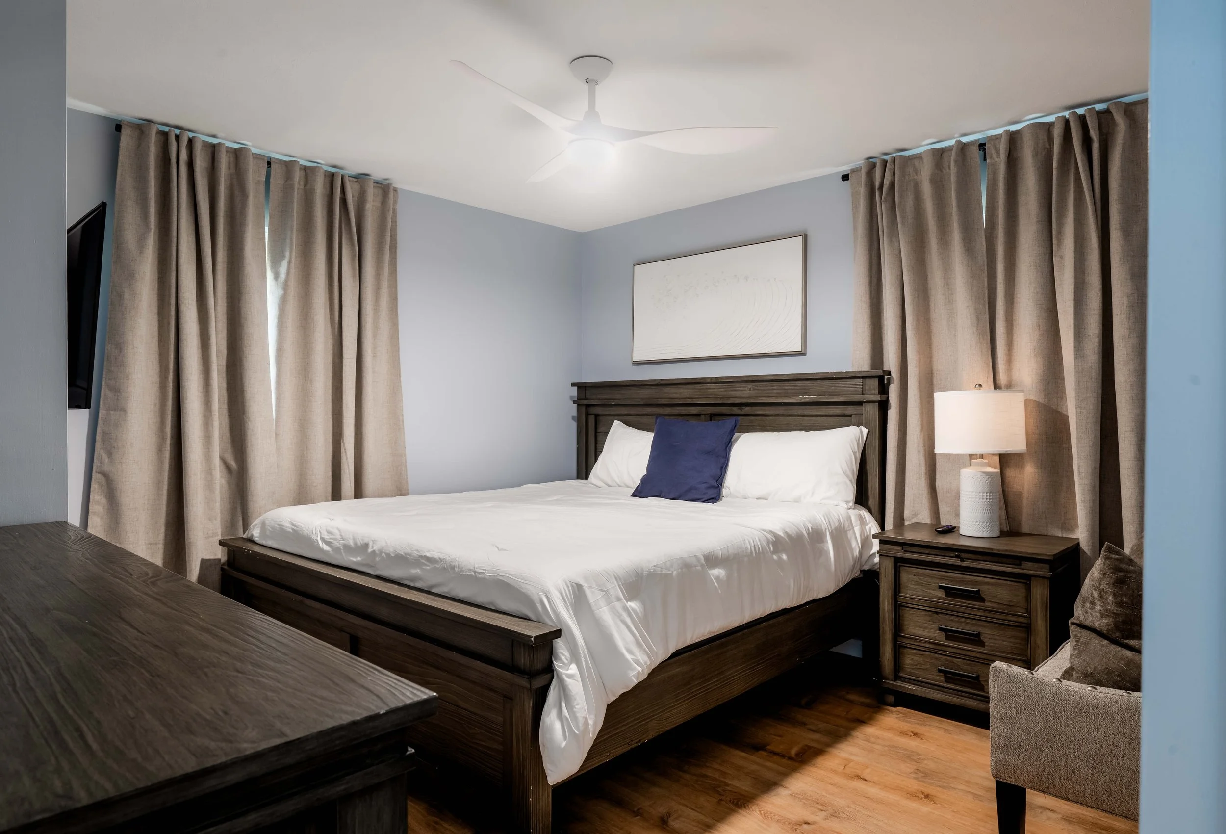 A bedroom with a wooden bed frame, white bedding, and a blue pillow. Beige curtains cover the windows, with a bedside table holding a white lamp and a remote. A framed artwork hangs above the bed, and a ceiling fan is installed on the white ceiling.