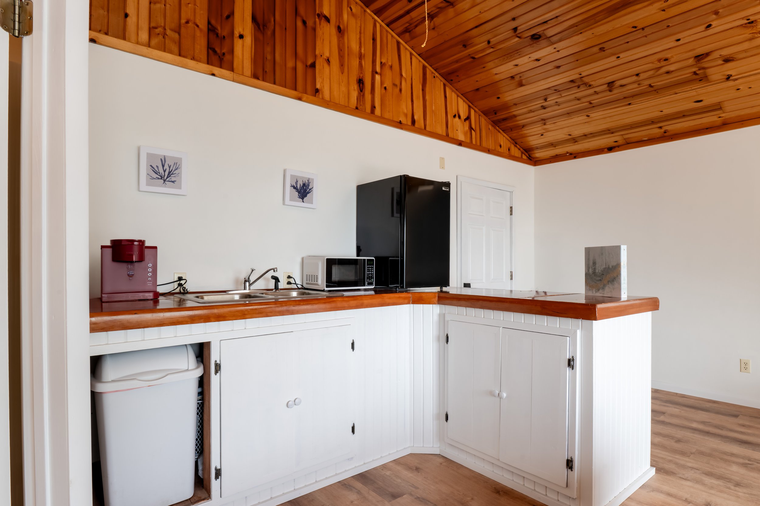 Kitchen area with white cabinets, countertop, black refrigerator, microwave, and red water dispenser on the counter. Two framed pictures of purple corals hang on the wall. Wooden ceiling and a partial view of a white door.