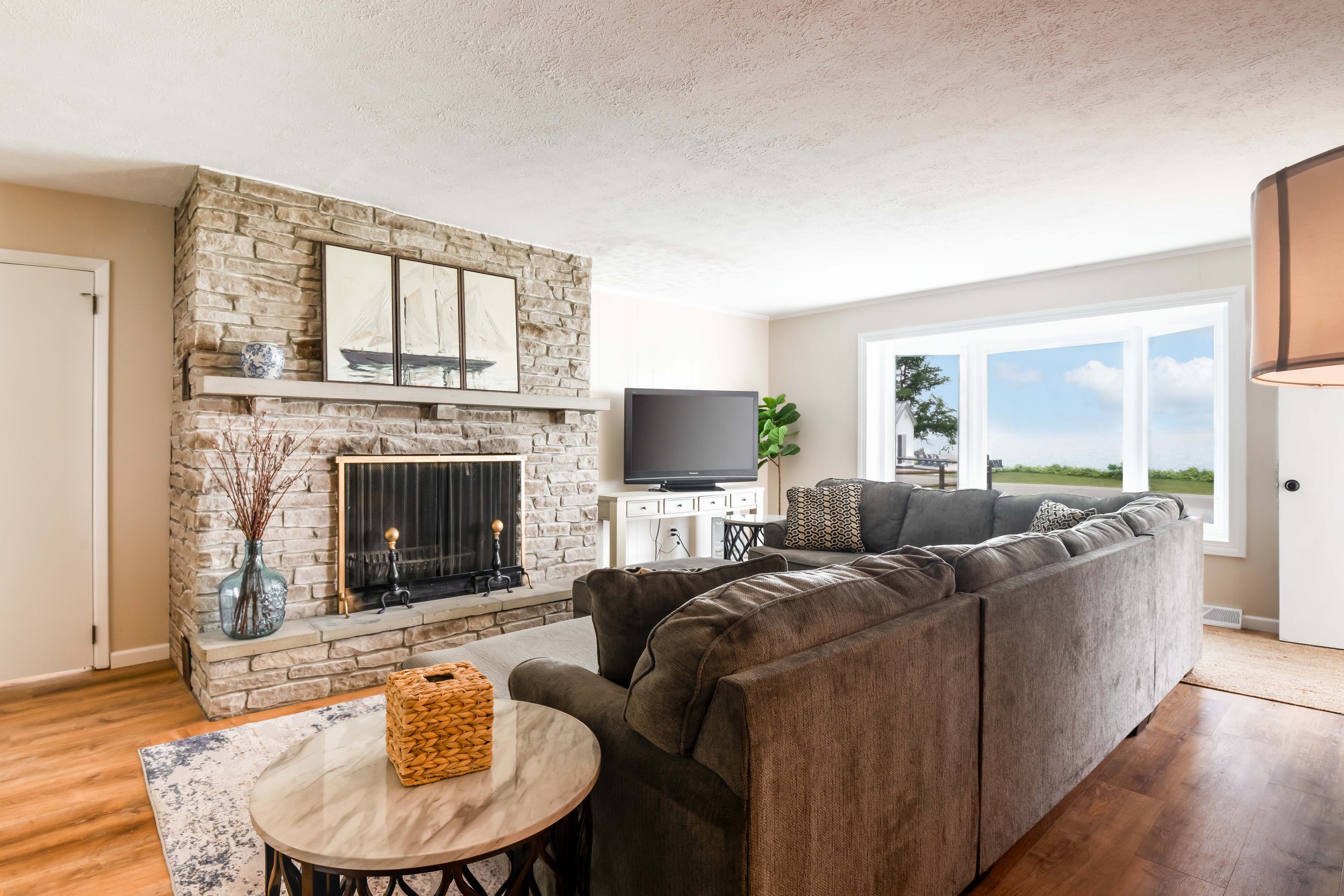 Living room with a stone fireplace, a flat-screen TV on a white stand, a sofa, a round marble coffee table with a woven basket, a large window showing a view of trees and sky, and indoor plants.