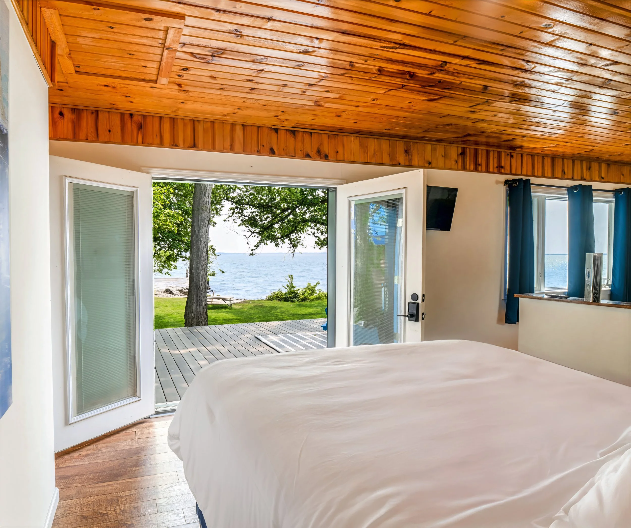 Bedroom with open glass door leading to a deck overlooking a lake, with trees and grass outside.