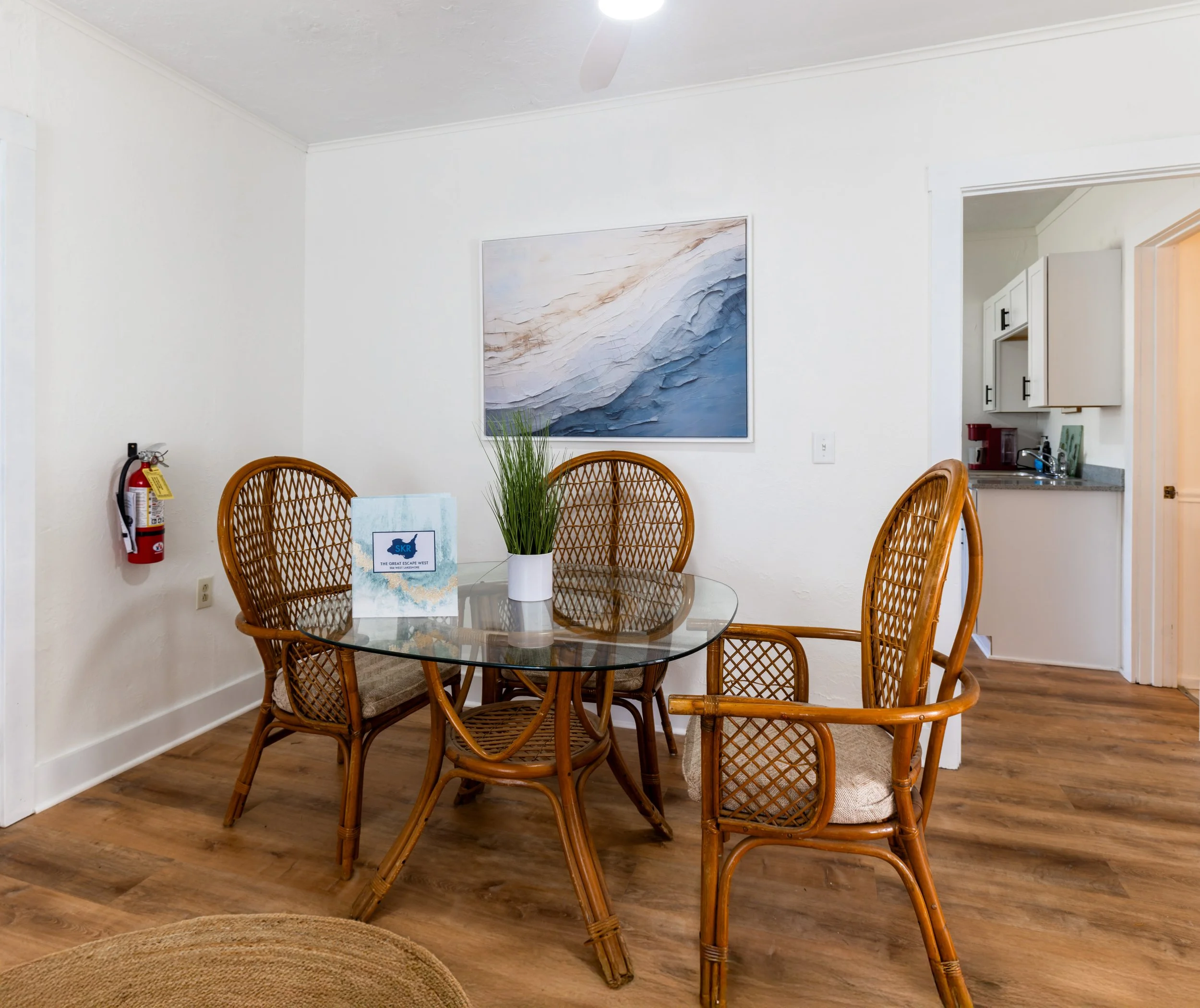 Dining area with wooden chairs around a glass table, a plant in a white pot, wall art, and view into kitchen with cabinets and a coffee maker.