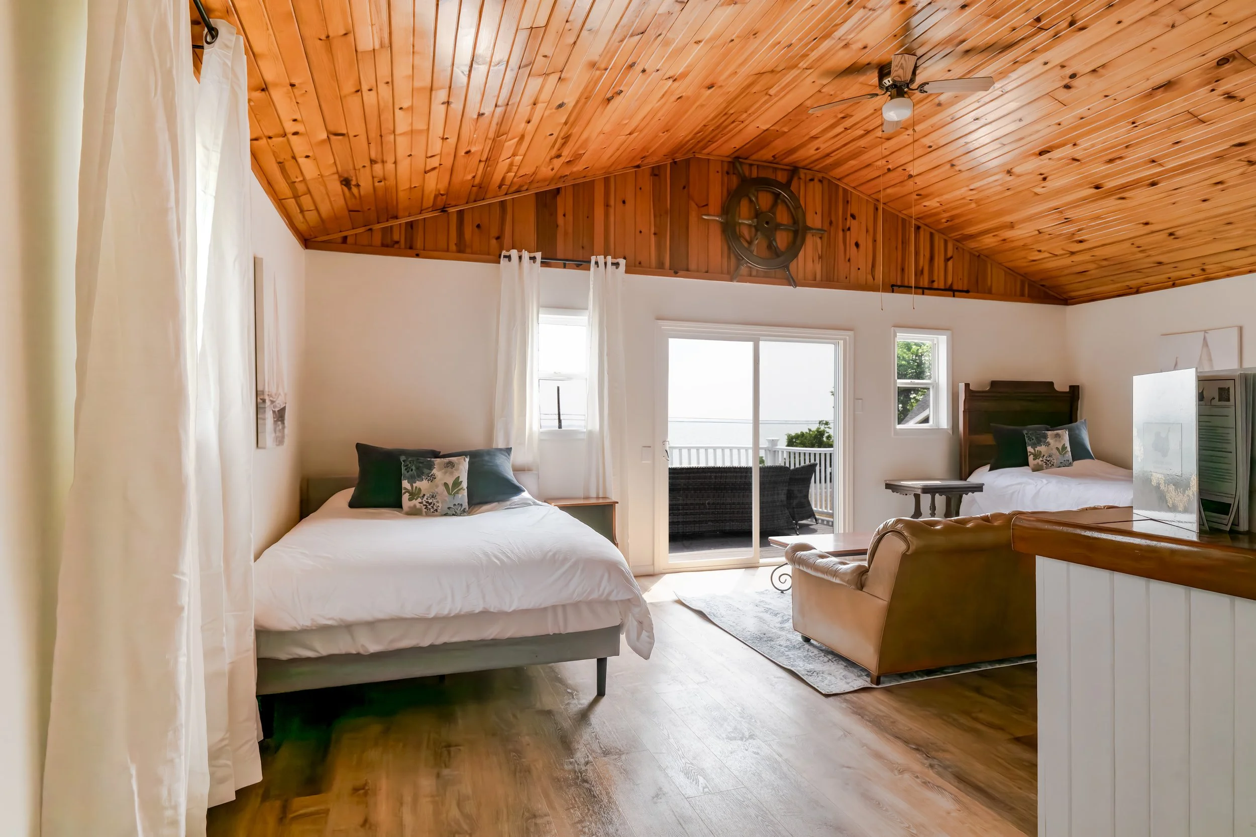 A cozy bedroom with a wooden vaulted ceiling and white walls. It has two beds with white bedding, decorative pillows, and a sliding glass door leading to a balcony with outdoor seating. The room features a ceiling fan, a wooden headboard, and large windows with white curtains.