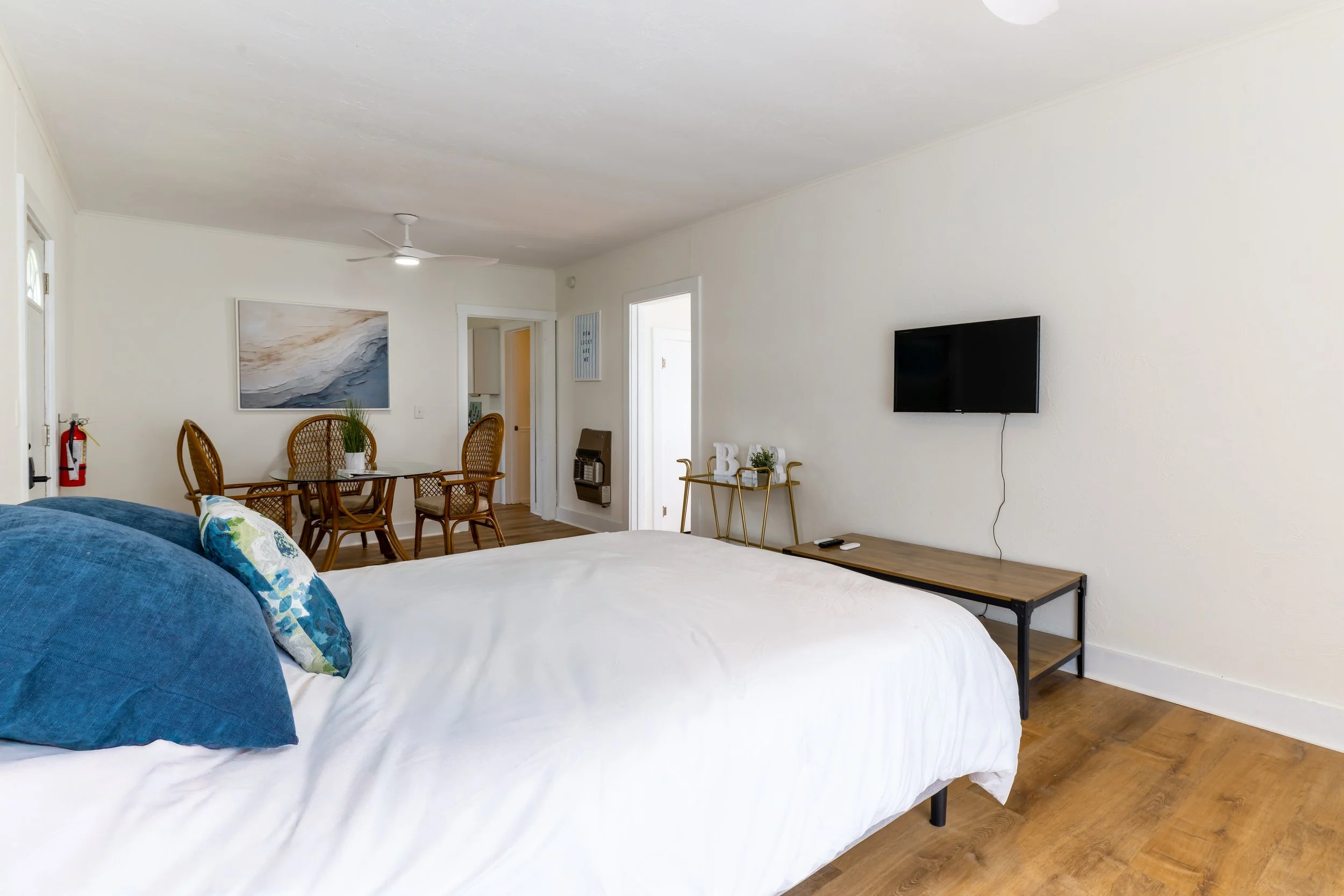 A bedroom with a bed in the foreground covered with white sheets and blue pillows, a wall-mounted TV, a wooden dining table with chairs, and minimalist decor.