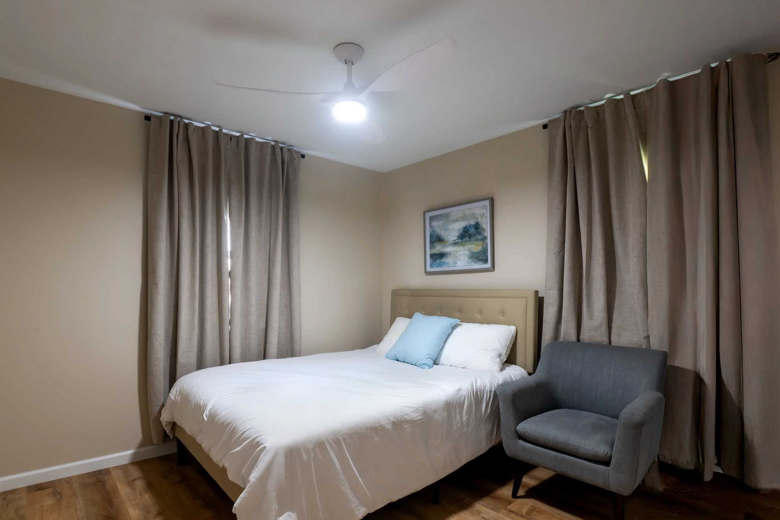 A bedroom with a bed, beige walls, curtains, a framed landscape painting, a ceiling fan with lights, and a gray armchair.