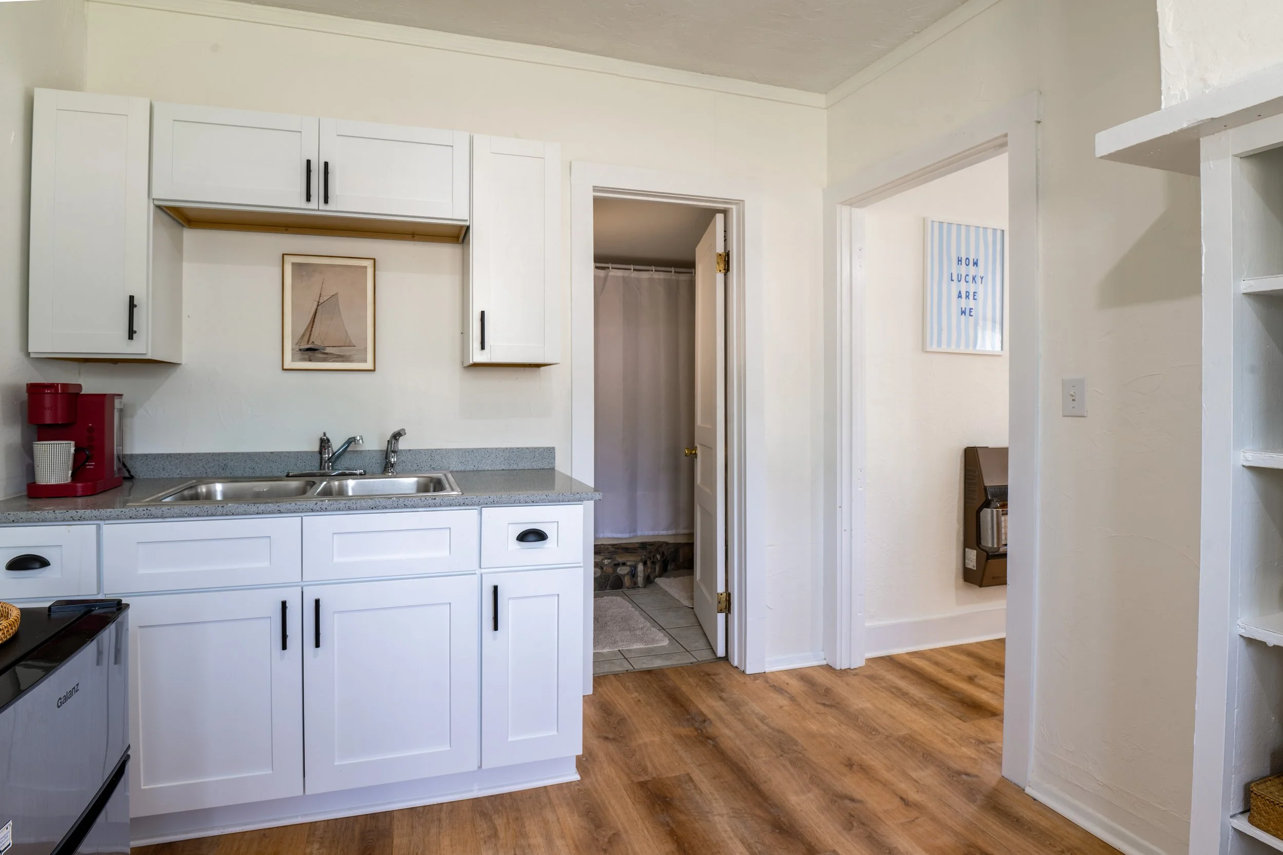 A kitchen with white cabinets and a gray countertop, a double sink, a small picture of a sailboat on the wall, and a doorway leading to a bathroom with a shower curtain. There is a red coffee machine on the counter and a wooden floor.
