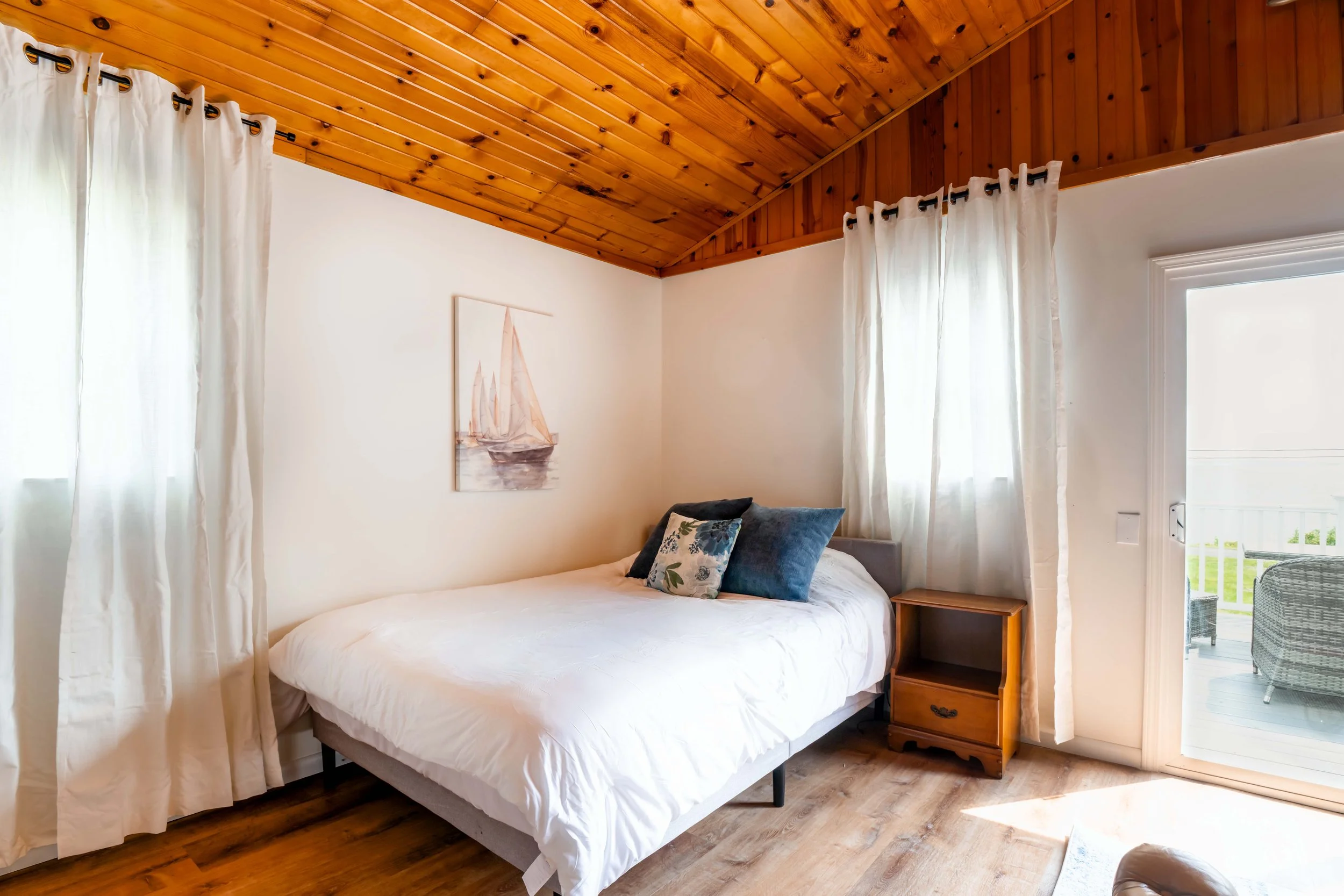 A bedroom with a wooden ceiling, white walls, and white curtains. There is a bed with pillows and a painting of a sailboat on the wall. A small wooden nightstand is beside the bed, and a glass door leads to an outdoor area.
