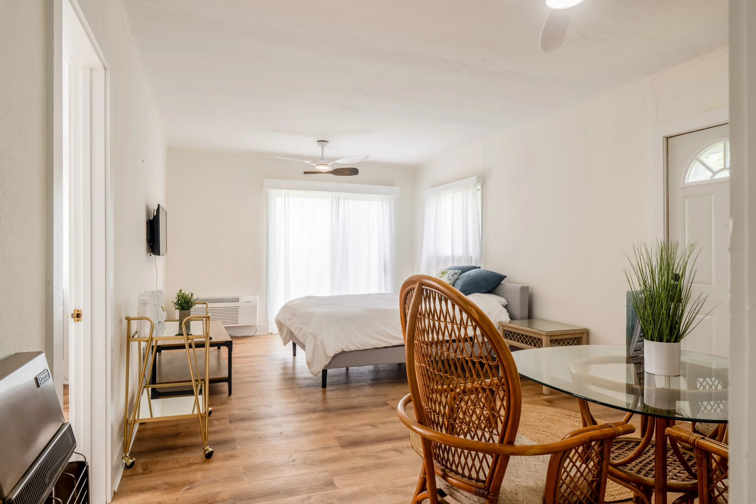 A bright living space with a bed, wicker chairs, a glass table, and large windows with white curtains, decorated with green plants and minimal furniture.