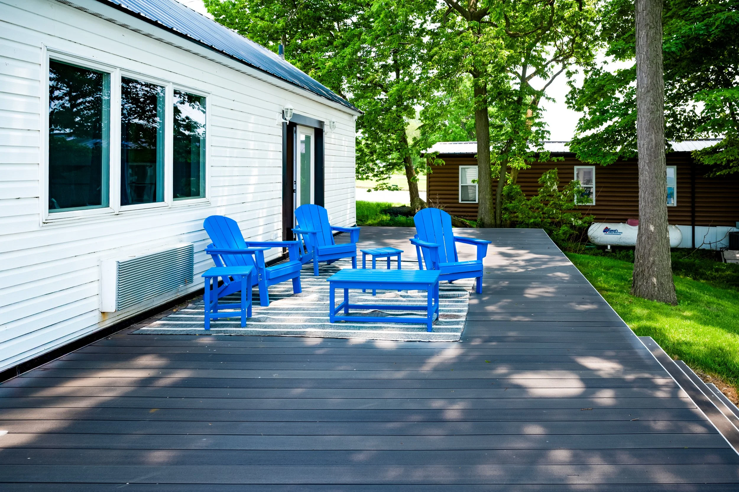 A backyard porch with four bright blue Adirondack chairs, a small matching table, and a patio rug. The porch is adjacent to a white house with horizontal siding, surrounded by green trees and a grassy yard.