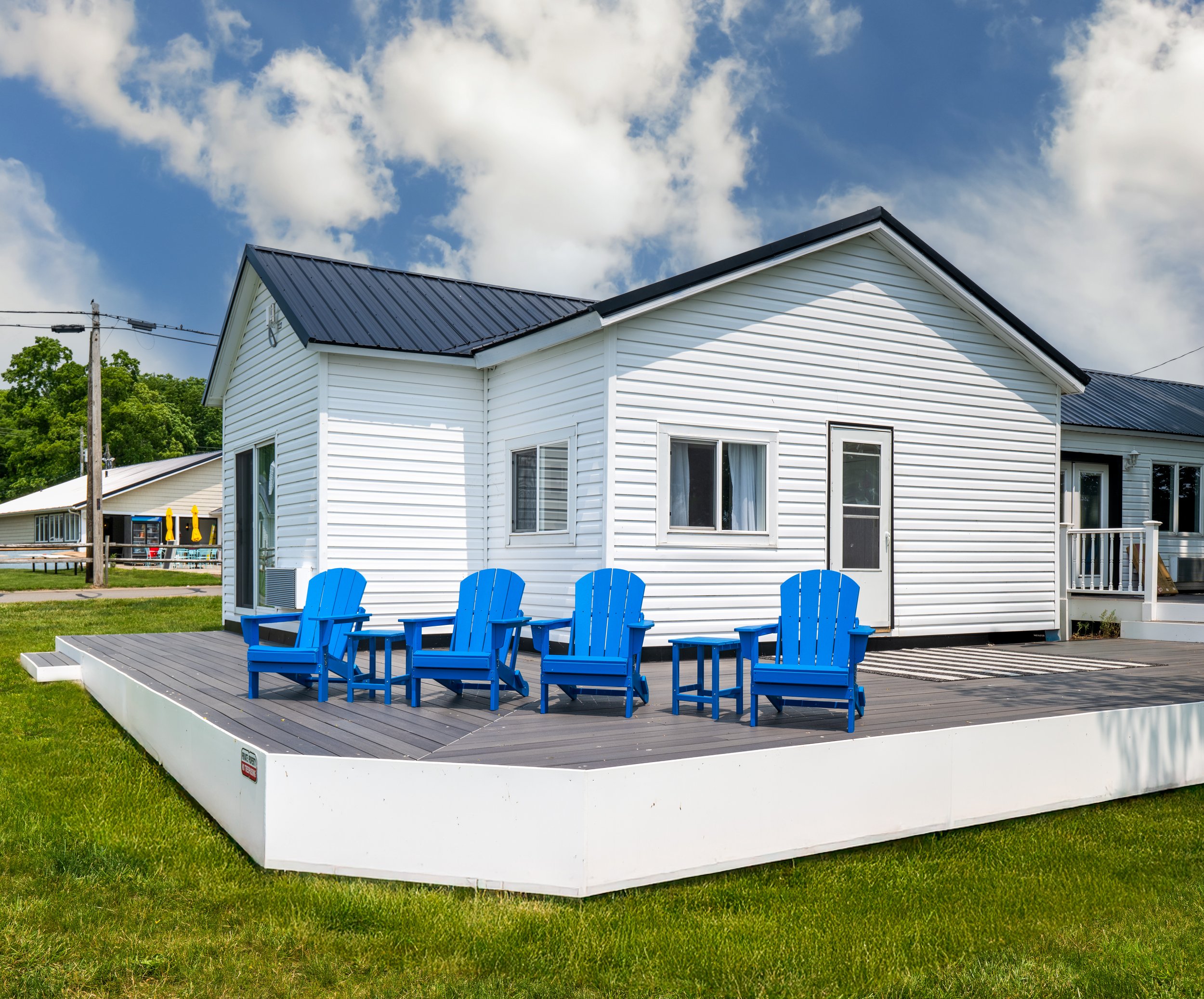 A white house with a black metal roof and several windows, set against a partly cloudy sky. In front of the house, there is a wooden deck with blue Adirondack chairs and a striped rug. Green grass surrounds the area.
