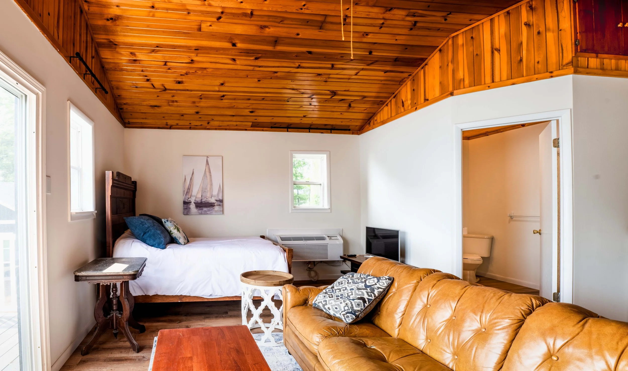 A cozy bedroom with a wooden vaulted ceiling, a bed with pillows, a small side table, a TV, and a bathroom visible through an open door.