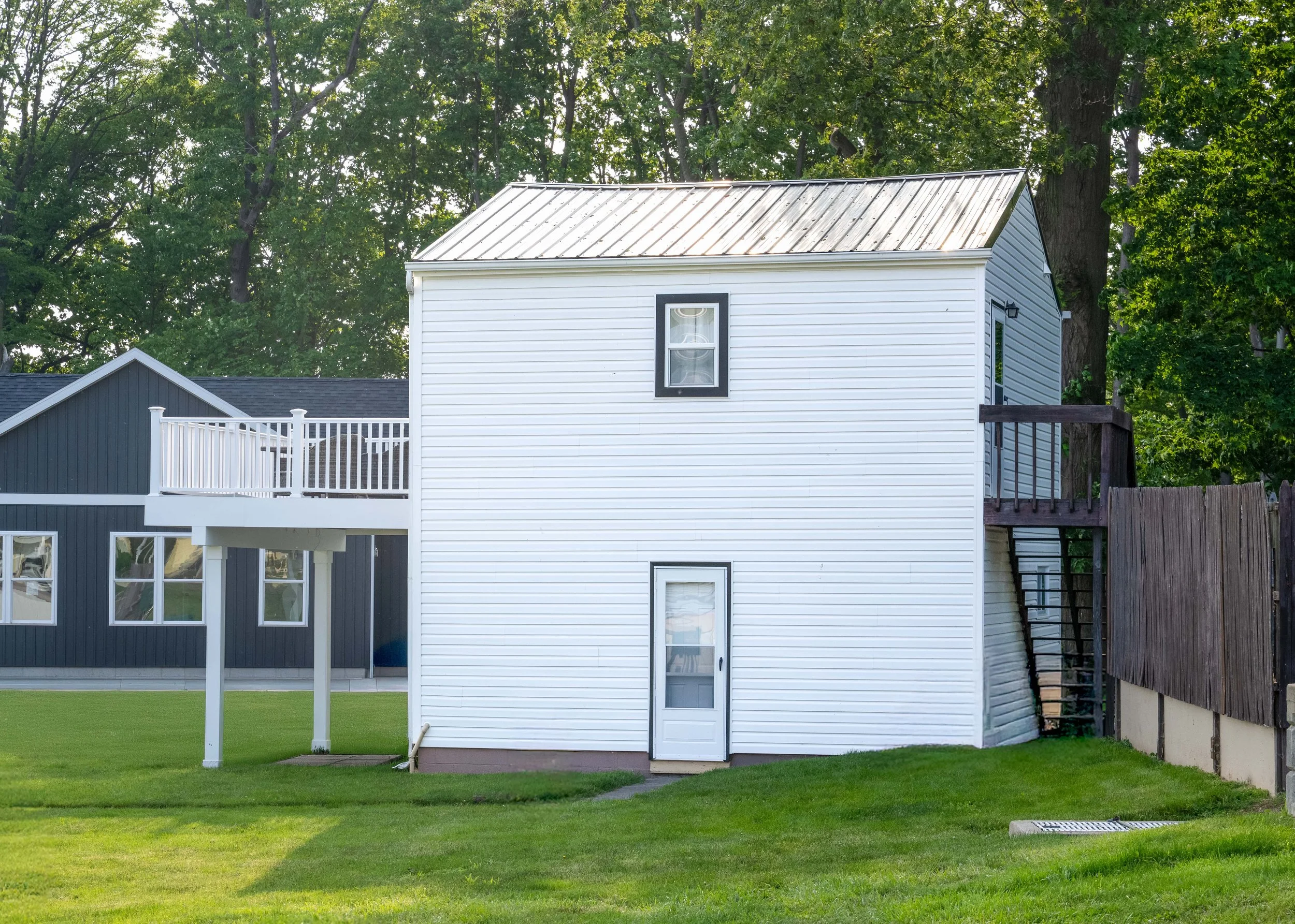 A backyard scene with a white multi-story house, green grass, and large trees in the background.