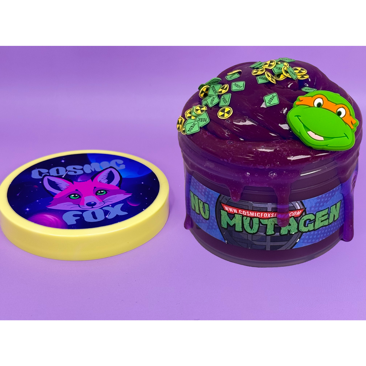 NU MUTAGEN Clear Based slime. Scented grape bubblegum and glows indigo in the light. Pictured with Special Edition Yellow Lid, TMNT Charms and clay sprinkles. Laid on a lavender background.