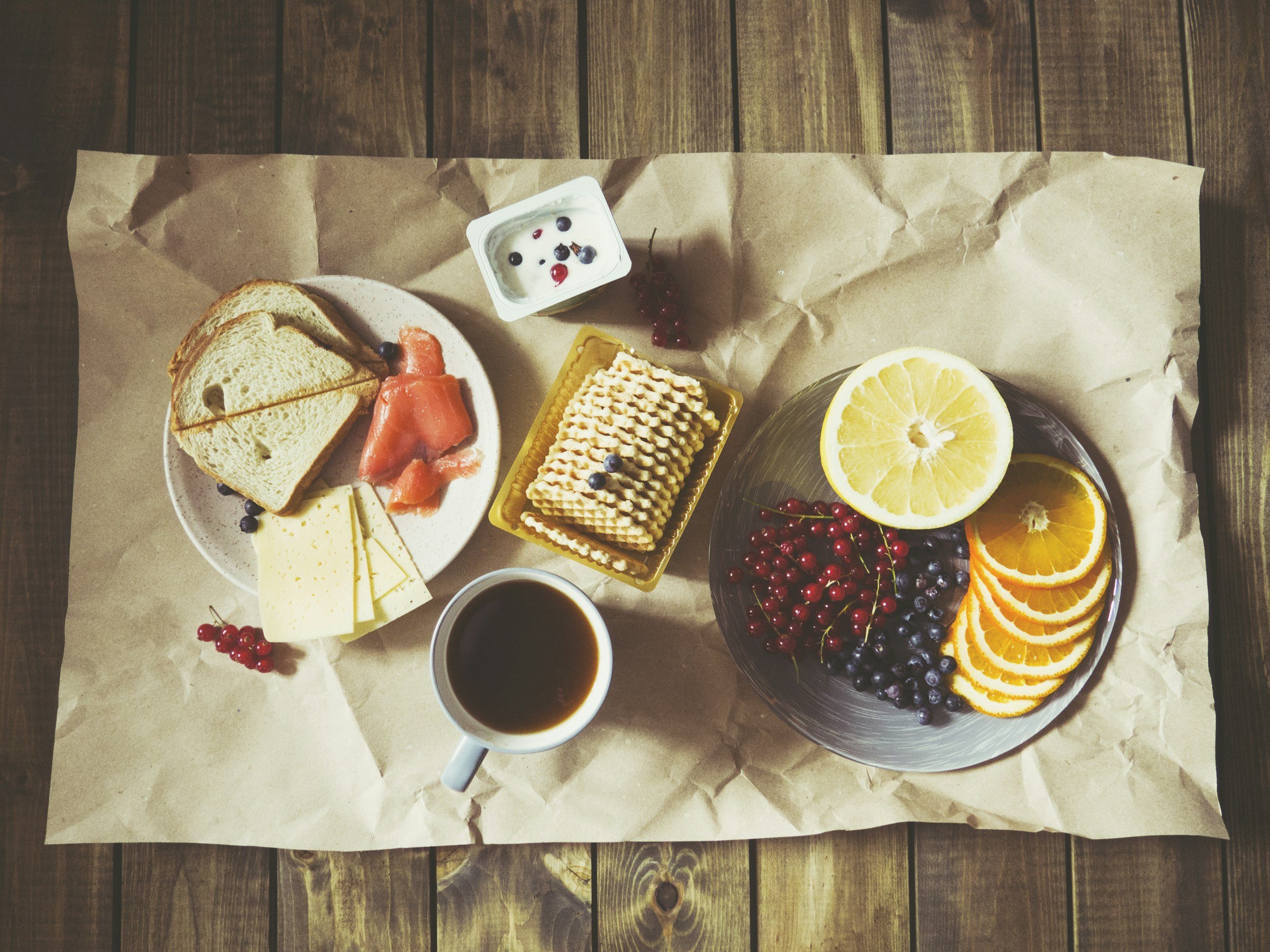 Breakfast spread on brown paper on a wooden table, includes sliced bread, smoked salmon, cheese, a small cup of yogurt with berries, a plate of sliced citrus fruits, a cup of coffee, and a small container of crispy wafers.