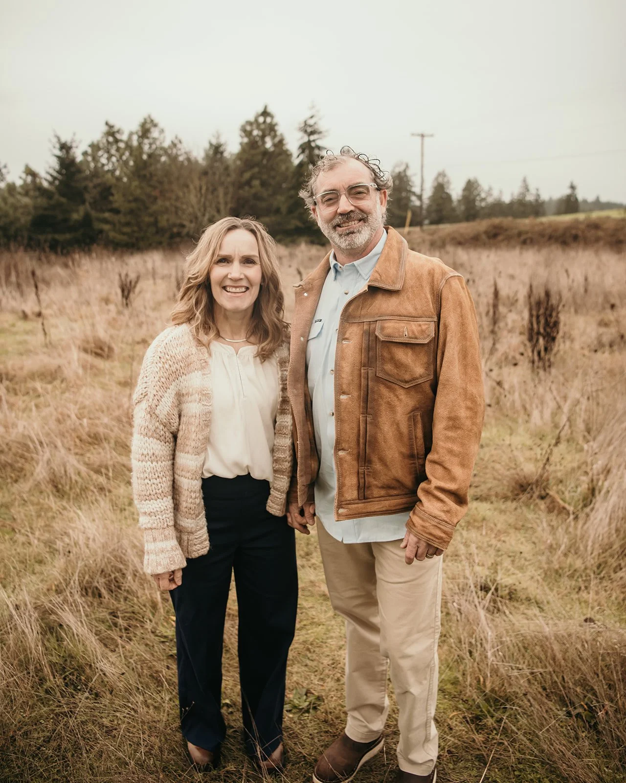 A smiling middle-aged couple holding hands outdoors in a grassy field with trees in the background.