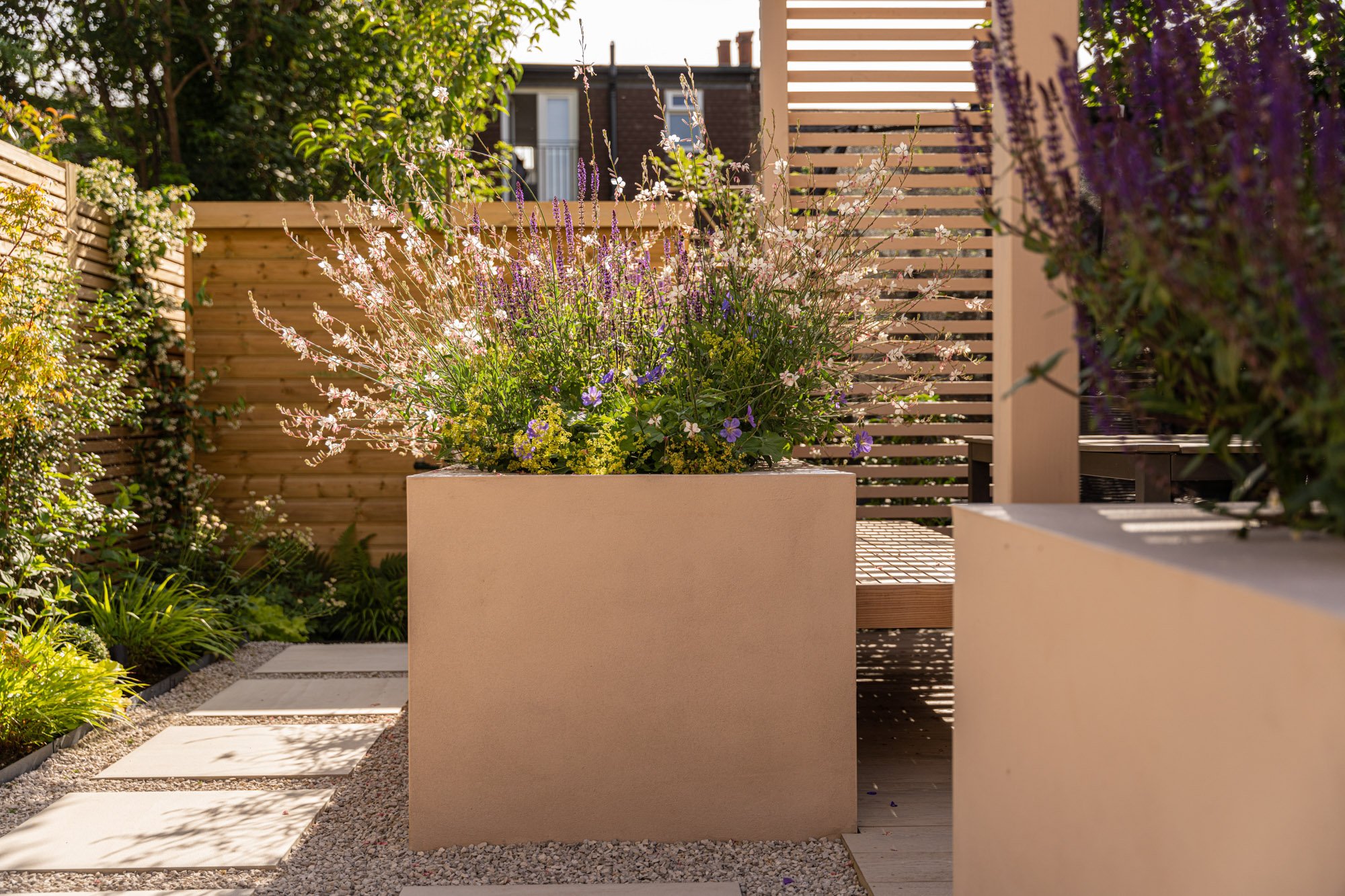 A modern outdoor garden scene with a large beige rectangular planter filled with flowering plants and purple flowers. The garden has a gravel pathway with square stepping stones, wooden fencing, and small trees with purple flowers on the right.