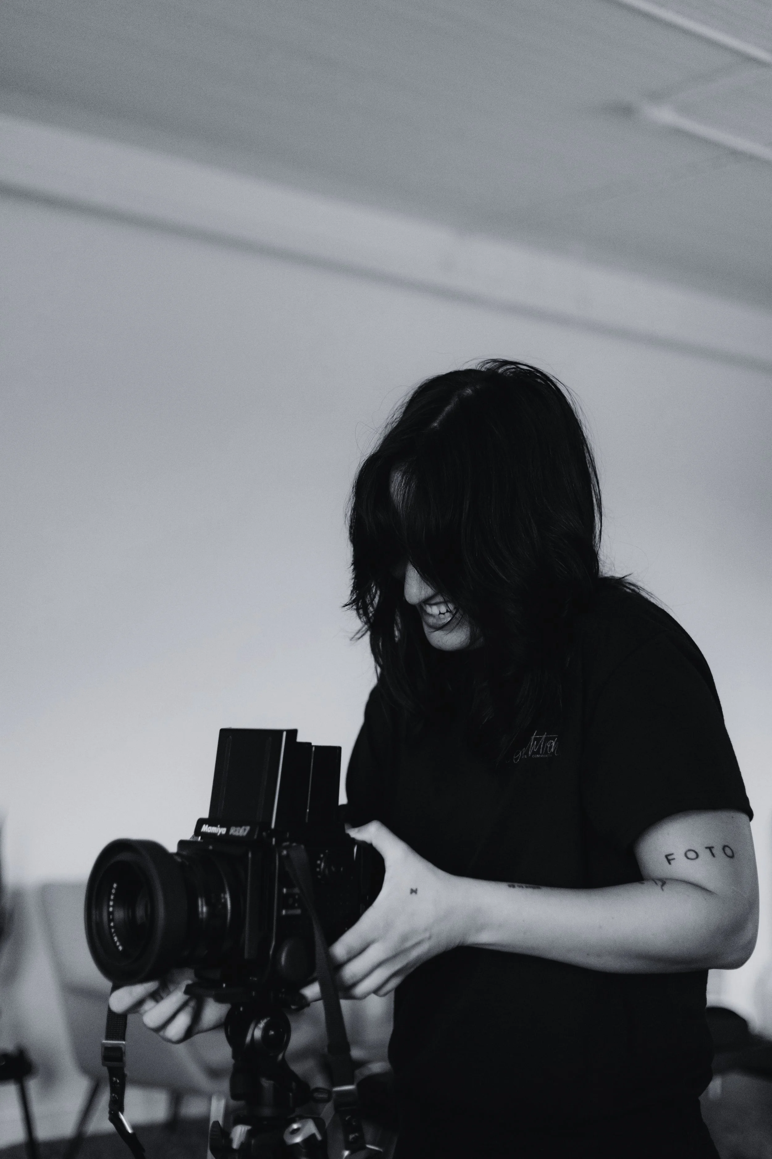 A female photographer with dark, wavy hair, smiling and looking down at a professional camera she is holding.