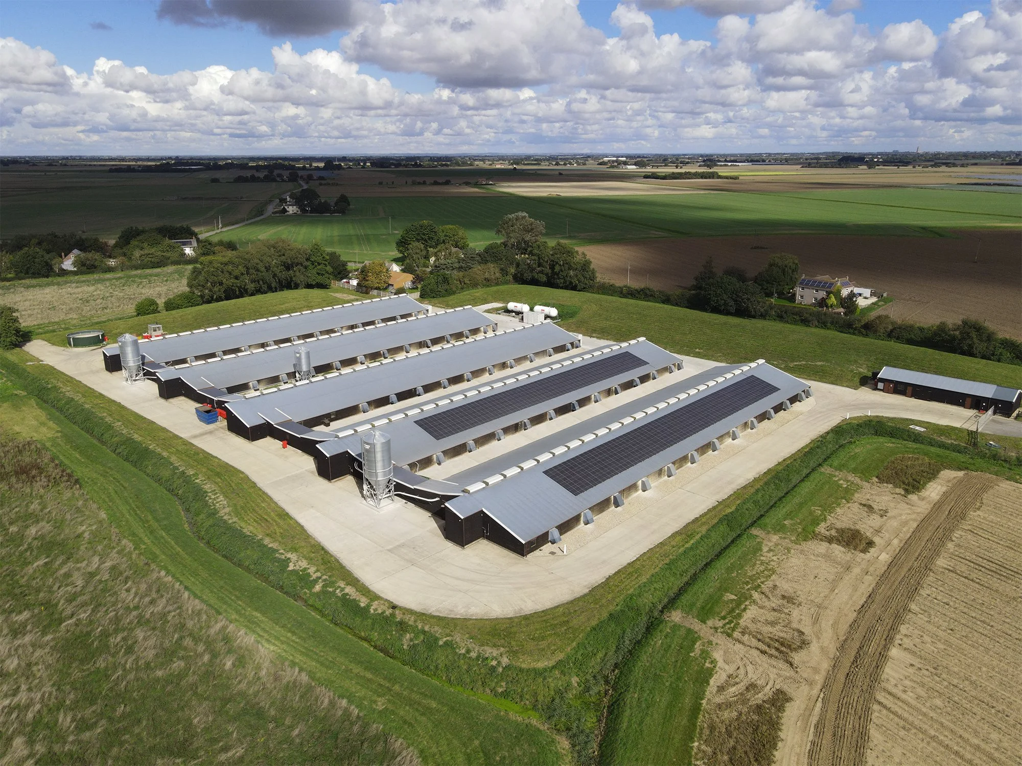 Aerial view of large agricultural buildings with solar panels on the roofs, surrounded by fields and grassland, under a cloudy sky.