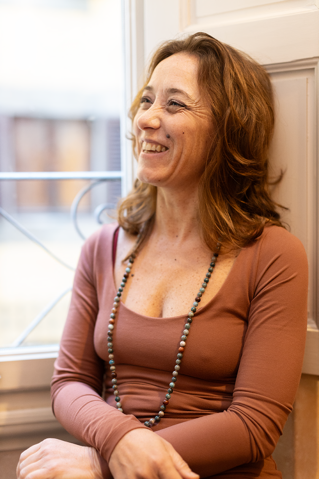 Elisa Batelli di Yoga e cambiamento, sorridente, seduta vicino a una finestra, indossa una maglietta marrone a maniche lunghe e una collana di perle multicolori.