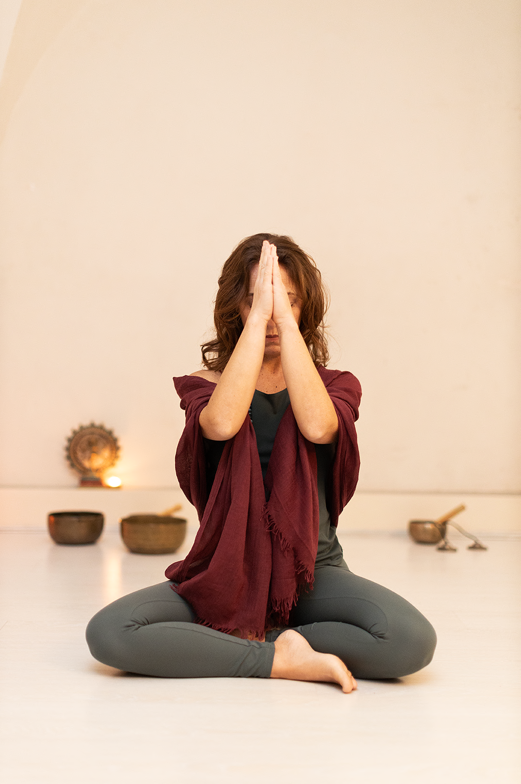 Una donna seduta a terra, con le gambe incrociate, in posizione di meditazione o preghiera, con le mani unite davanti al viso in segno di rispetto o preghiera, in un ambiente tranquillo con strumenti musicali sullo sfondo.