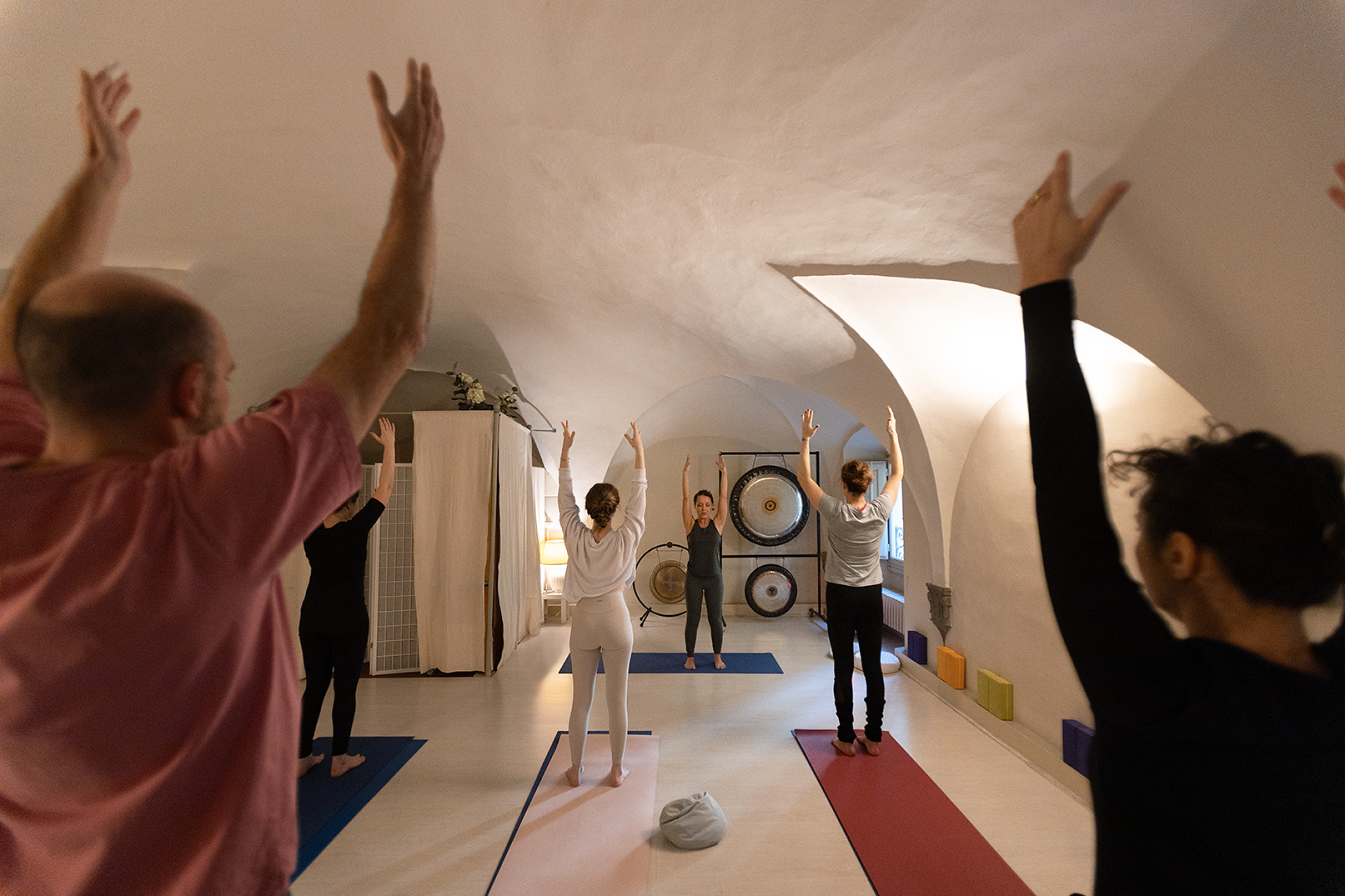 Gruppo di persone che praticano yoga in una stanza con soffitto arcuato, alcune con braccia alzate, su tappetini colorati.