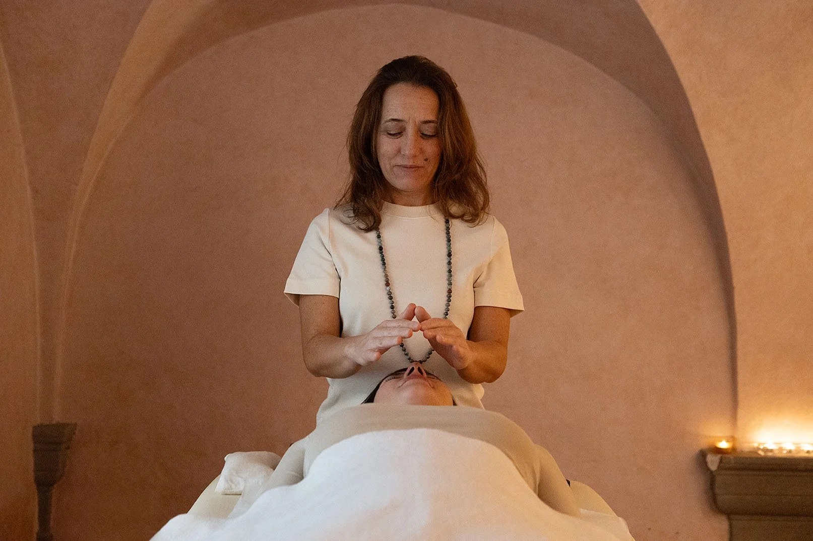 Una donna che riceve un trattamento di Reiki, con le mani sopra il viso di una persona distesa su un letto, in una stanza con pareti di colore caldo e luci soffuse.